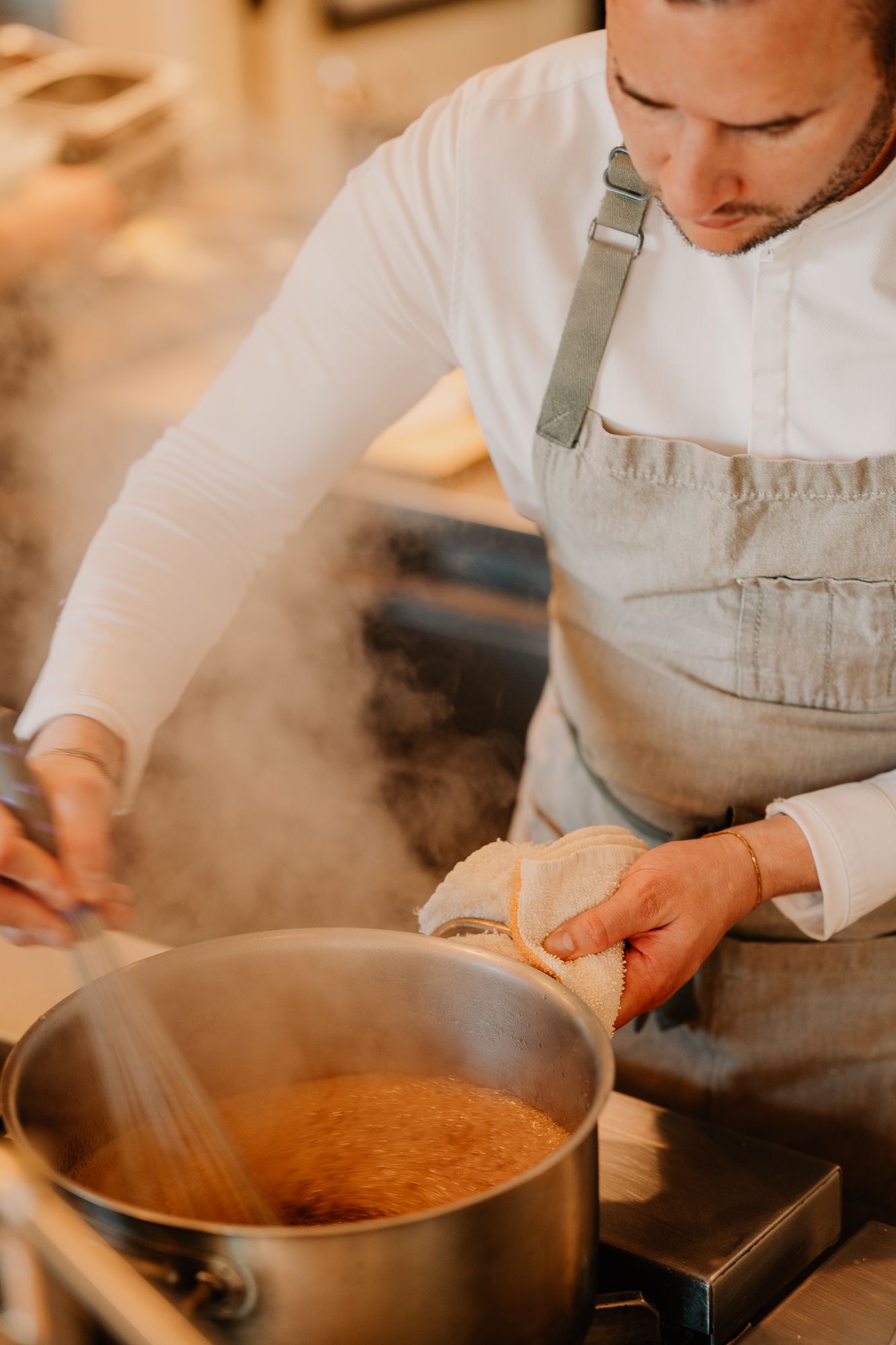 Chef in apron stirring steaming pot of sauce in professional kitchen.