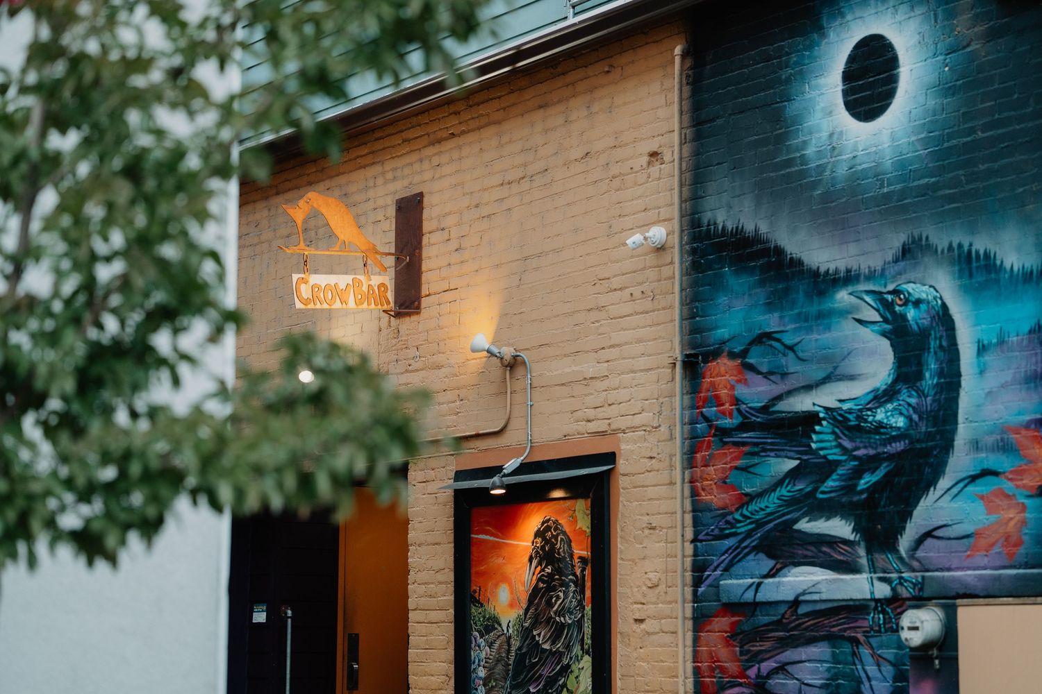 Street art mural of a raven against a moonlit sky adorns the brick wall of a building with an illuminated orange sign.