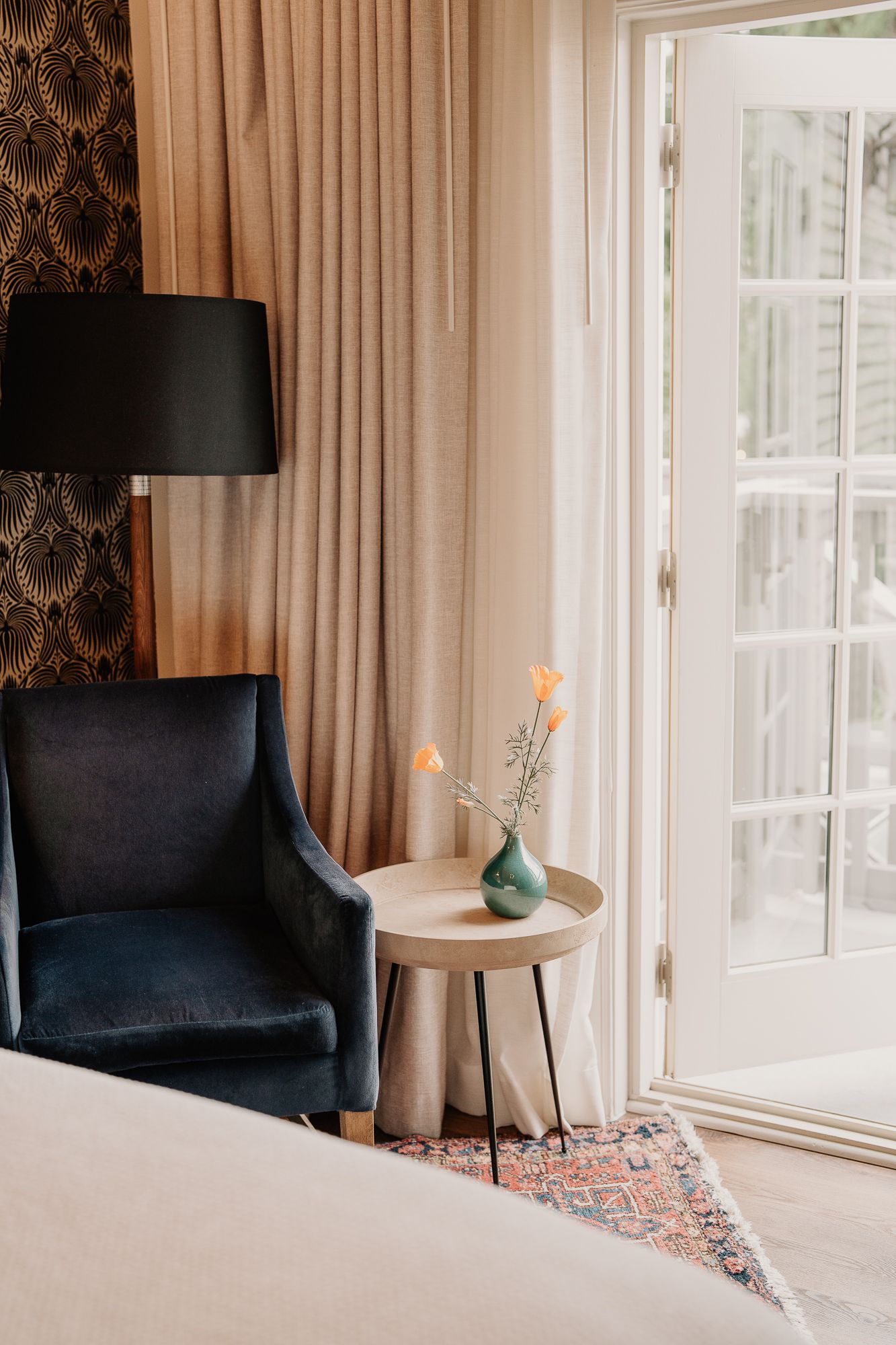 Modern bedroom interior with black accent chair, patterned wallpaper, and large windows with flowing beige curtains.