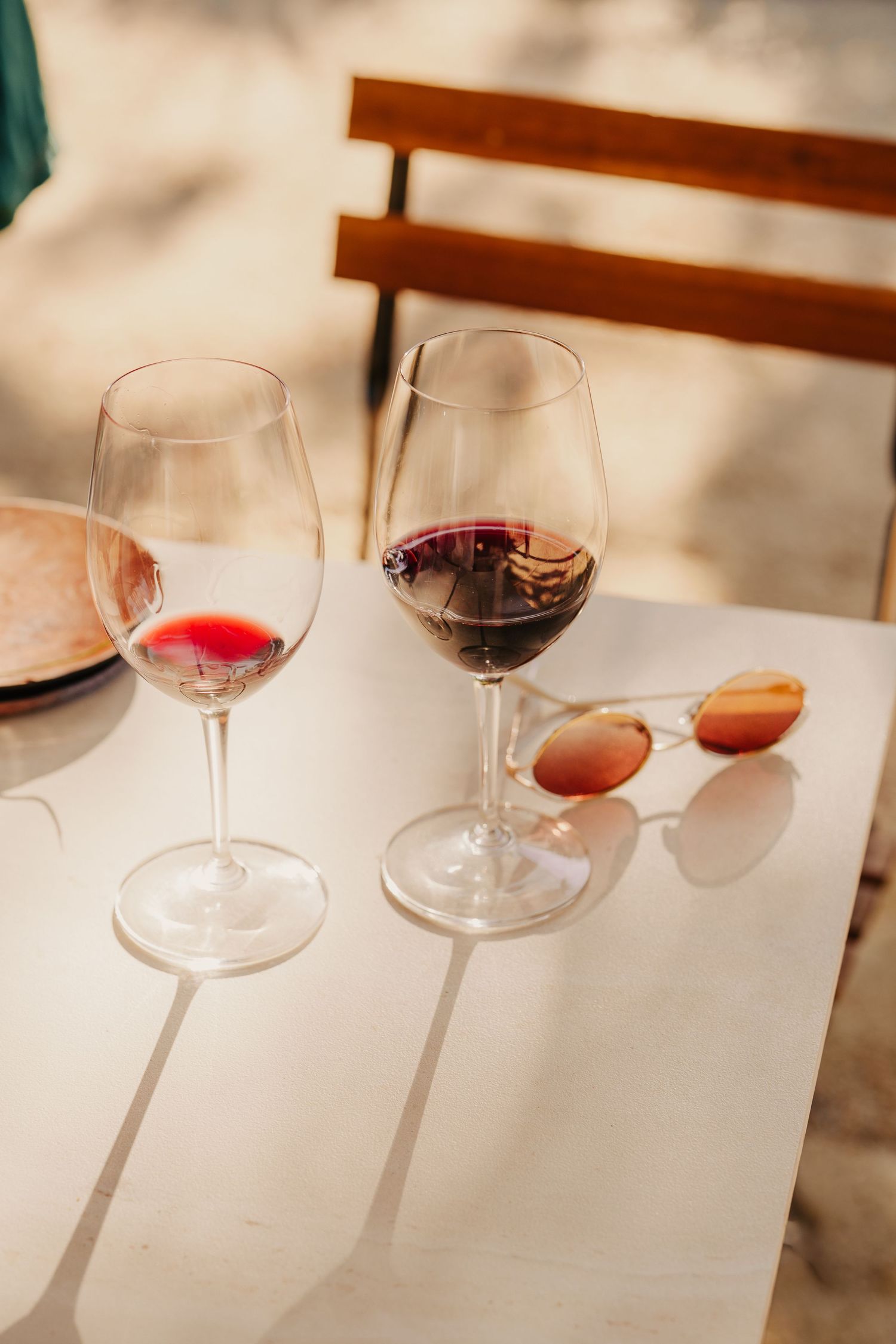Two glasses of red wine and sunglasses on a white marble table in warm sunlight outdoors.