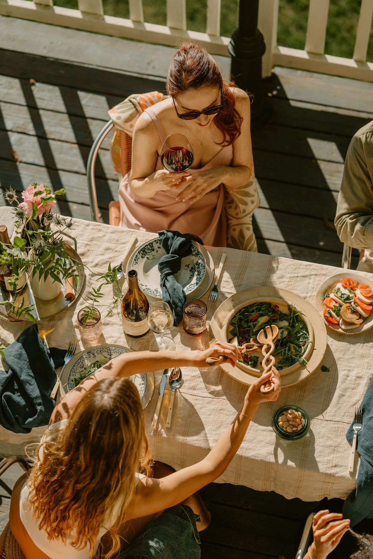 Two women dining al fresco on a sunny deck, sharing food and wine at a table with fresh flowers and dishes.