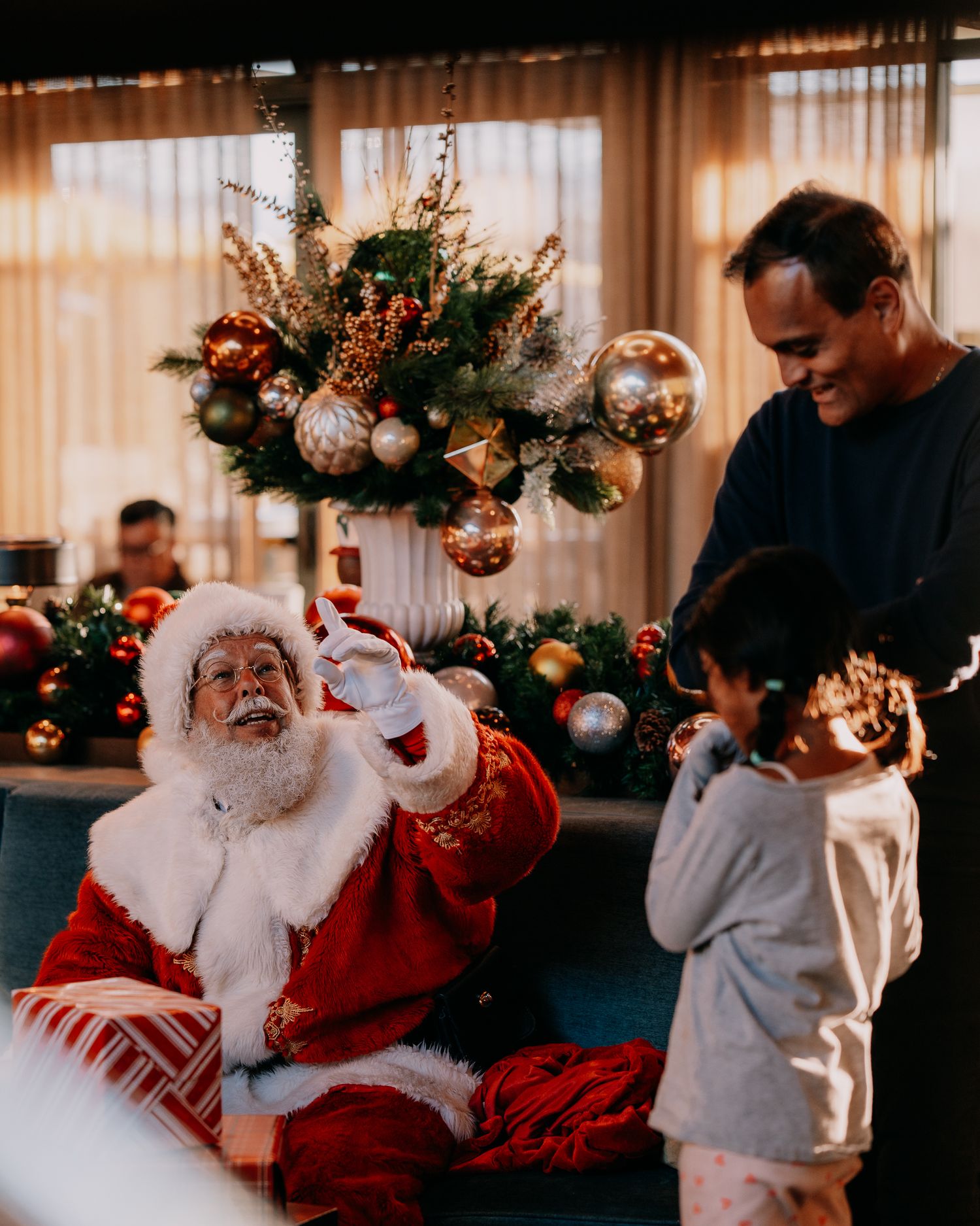 A festive holiday scene shows a person in a red Santa suit interacting joyfully with a small child in a grey sweater.