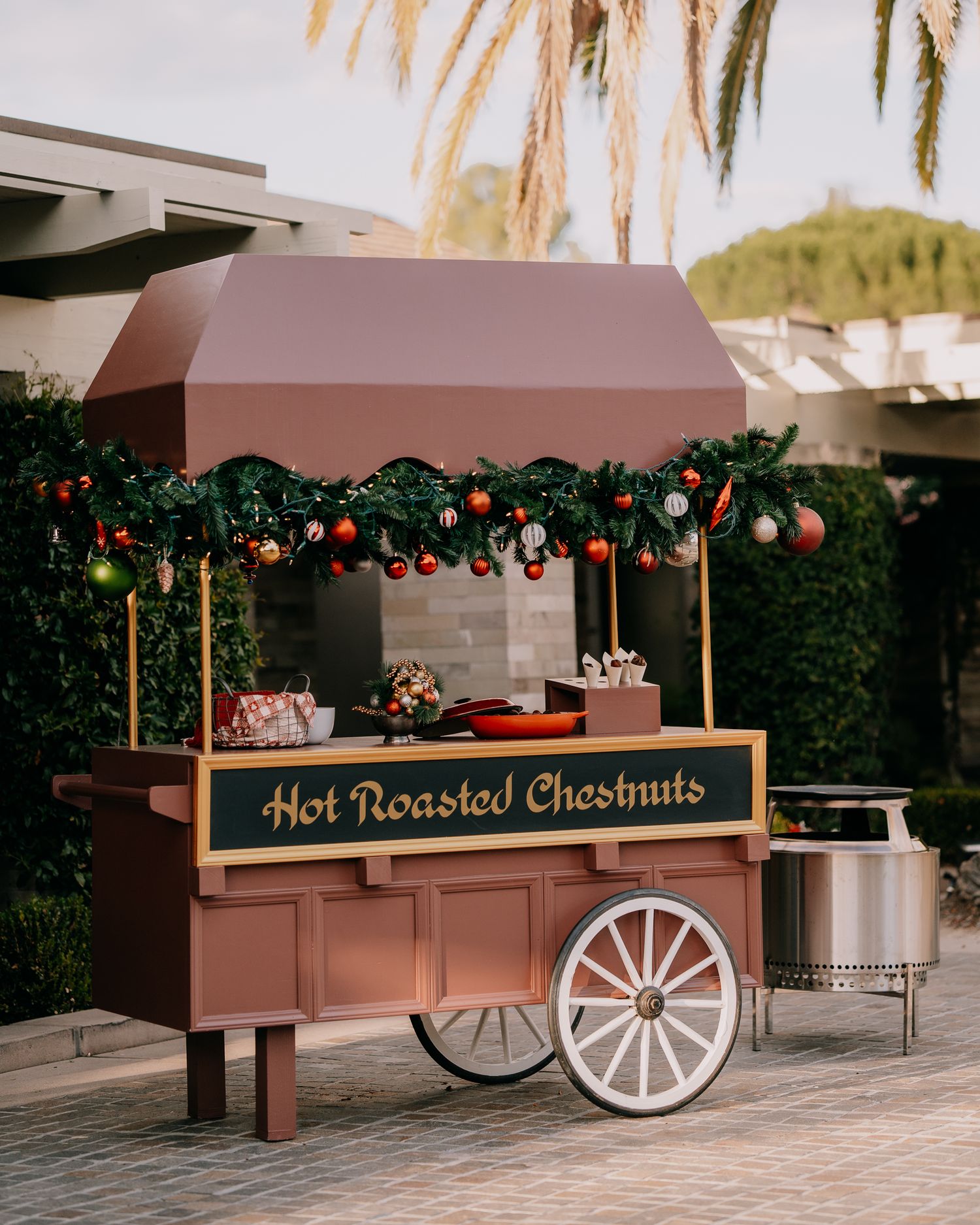 Wooden vendor cart with 'Hot Roasted Chestnuts' sign decorated with Christmas garland and lights at an outdoor market.