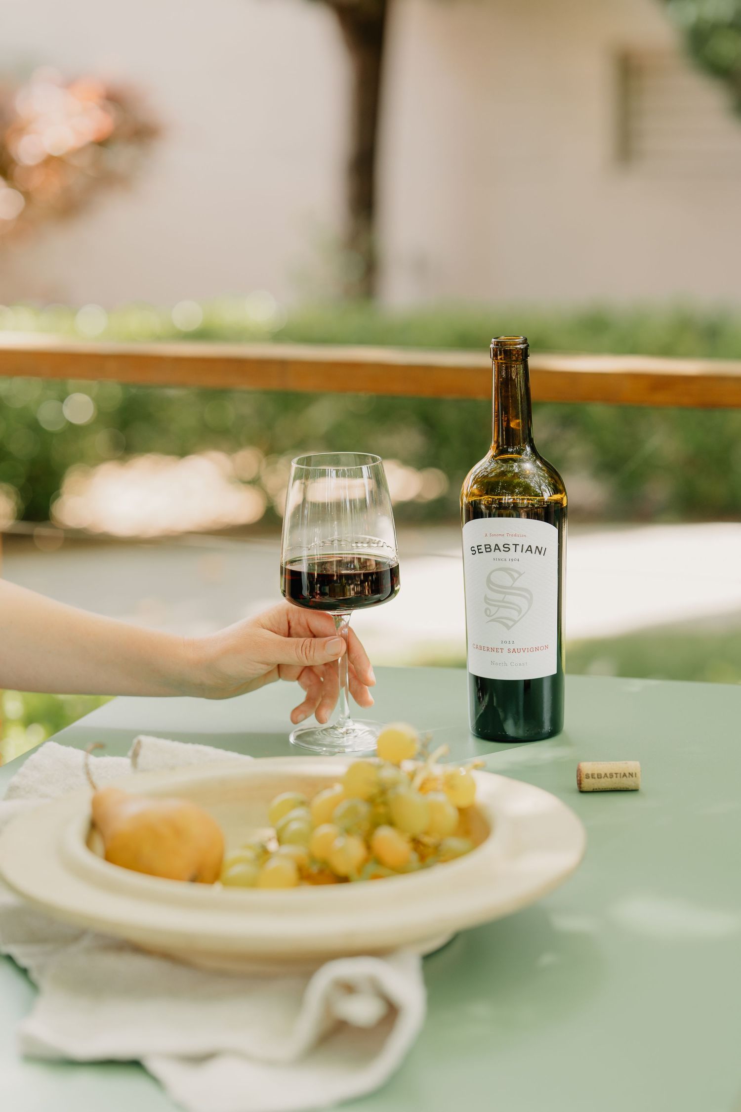 Hand holding Sebastiani Cabernet Sauvignon red wine glass beside fruit plate and bottle on outdoor table.