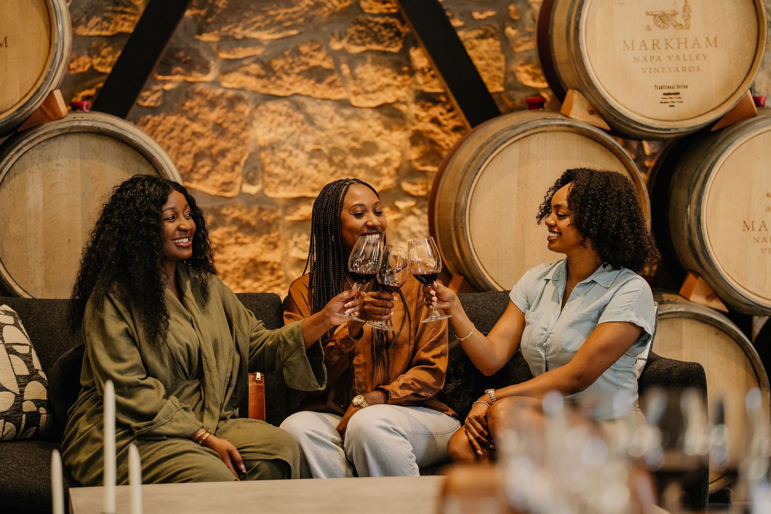 Three women smiling and toasting with red wine glasses in a rustic winery barrel room.