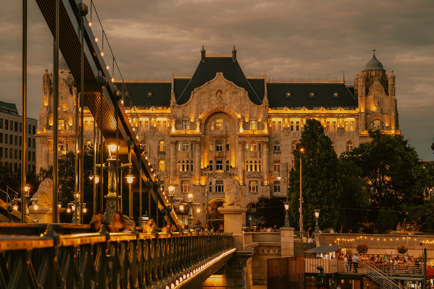 Illuminated historic building with ornate facade glowing at dusk, viewed from a suspension bridge in Budapest, Hungary.