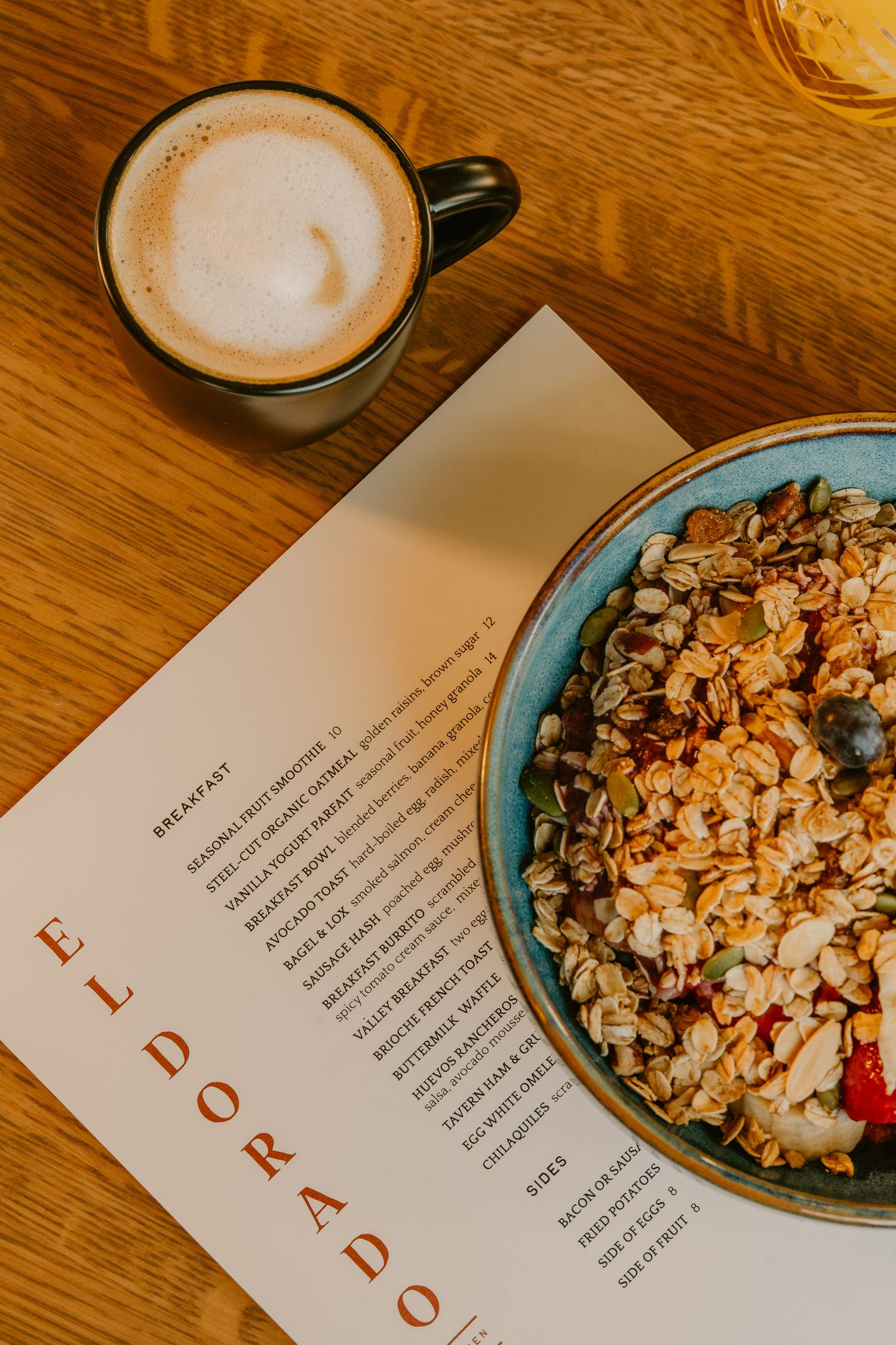 Coffee cups and granola bowls arranged with text documents on a wooden table in warm lighting.