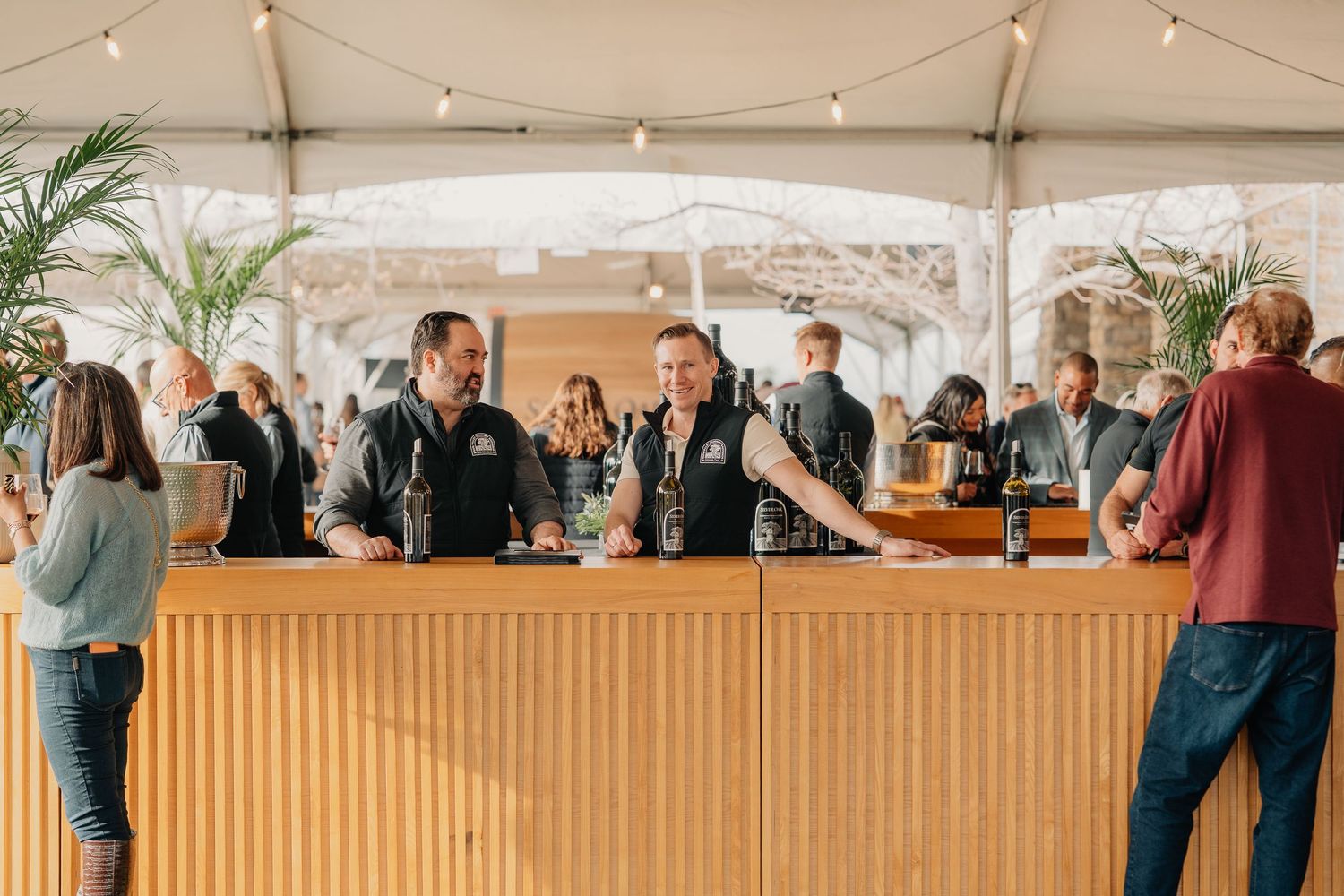 Two smiling staff members serving wine at a wooden bar counter inside a tent at an elegant outdoor event.