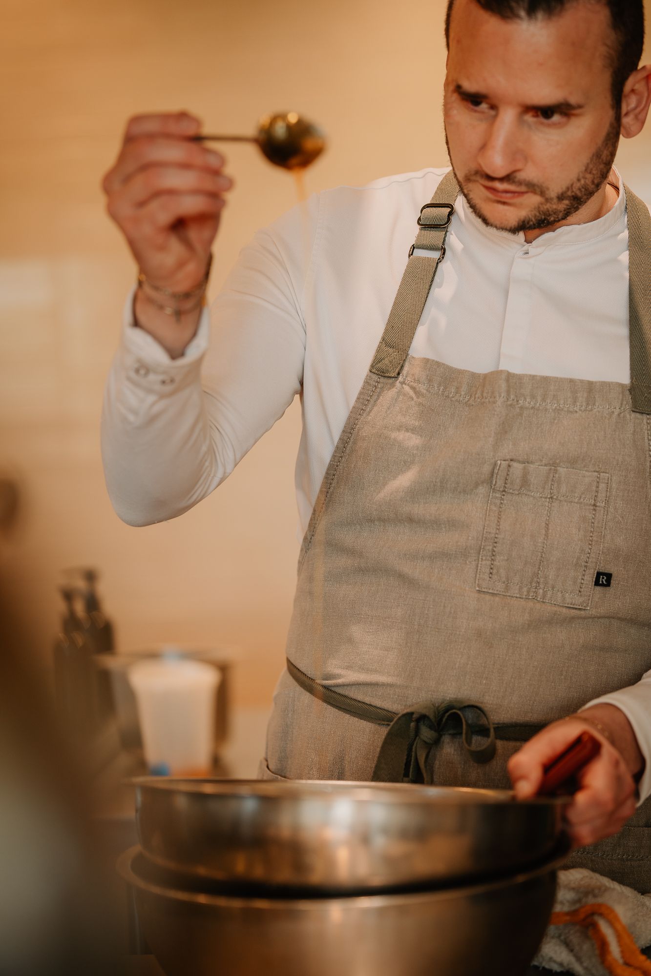 Chef in apron holding a ladle over a steel mixing bowl while cooking in a professional kitchen.