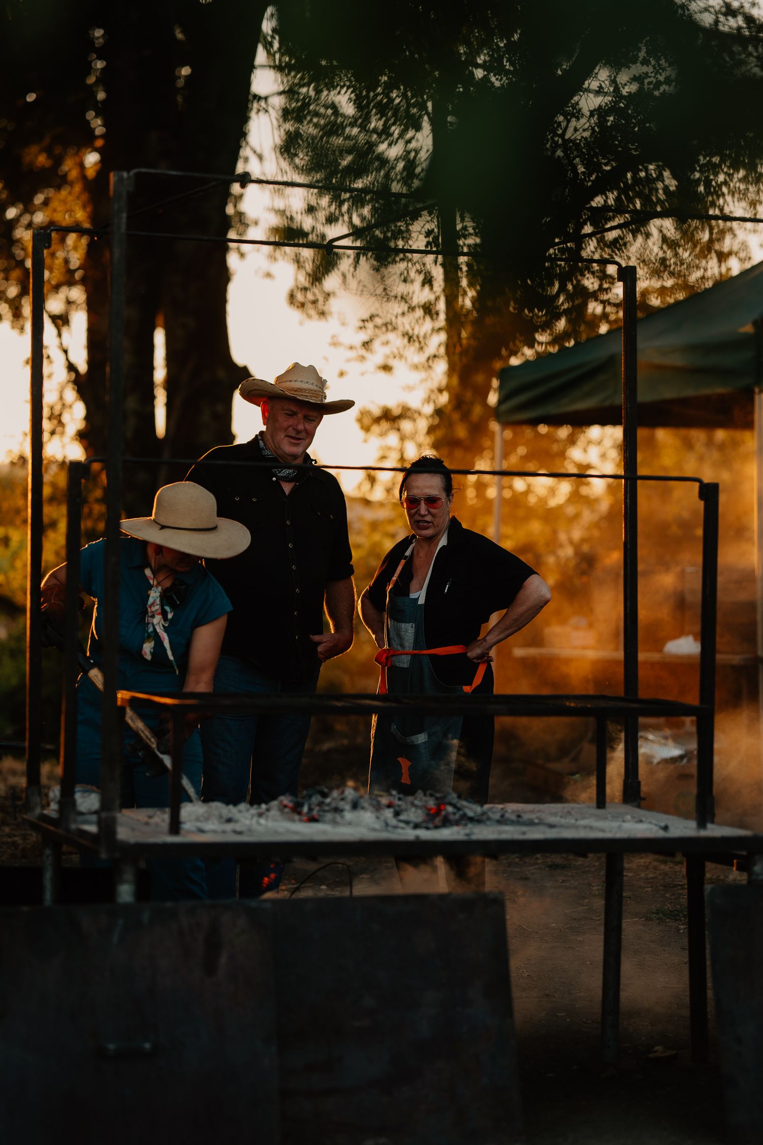Three people in cowboy hats gathered at a outdoor stage during golden sunset with atmospheric backlit lighting.