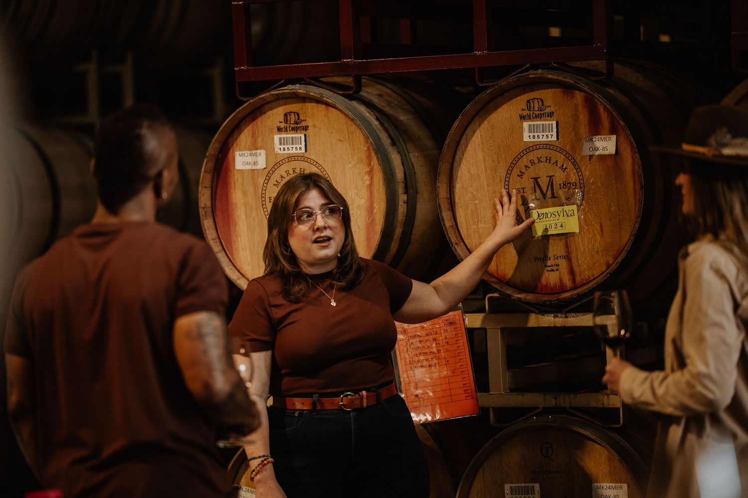 A woman gestures toward oak wine barrels in a dimly lit winery cellar during a barrel tasting tour.
