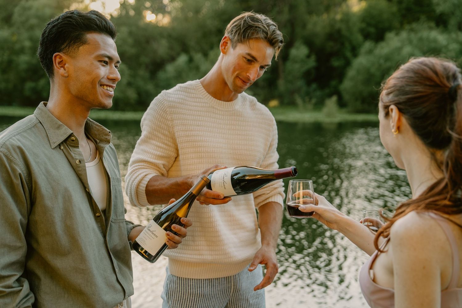 Three friends celebrating outdoors near a lake, with a man pouring champagne into a glass on a sunny day.