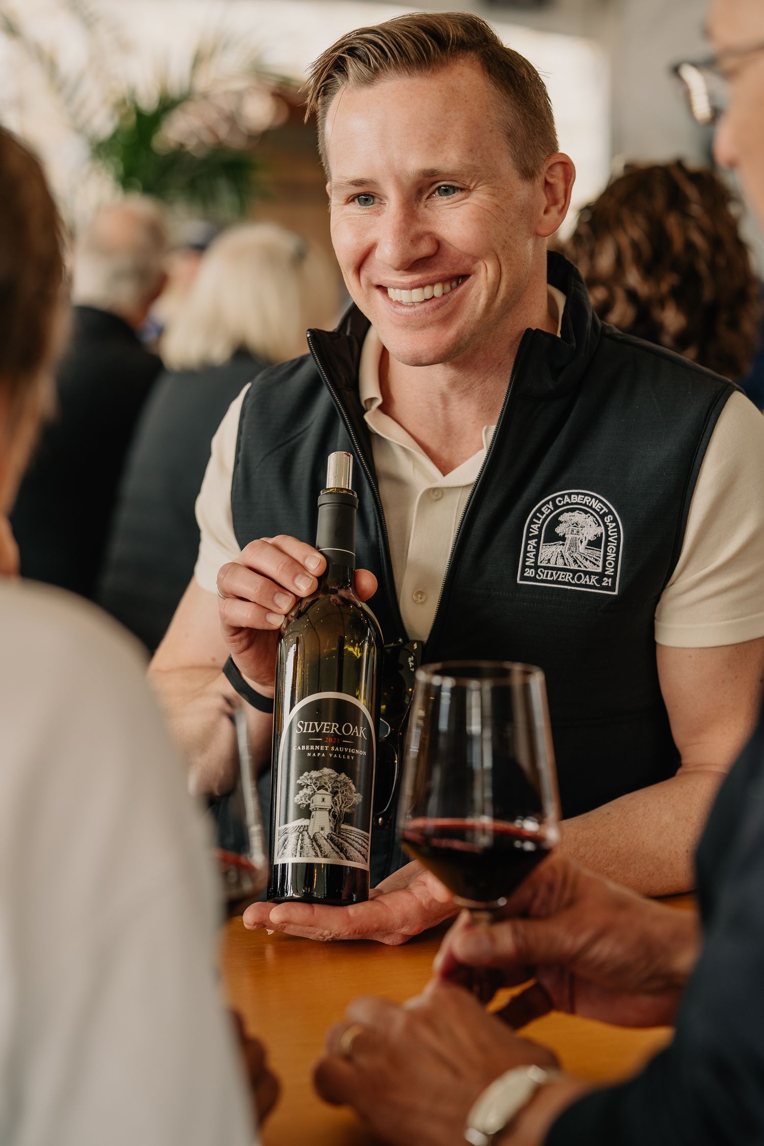 Smiling man in Silver Oak vest holding a bottle of Silver Oak wine at a tasting event with guests.