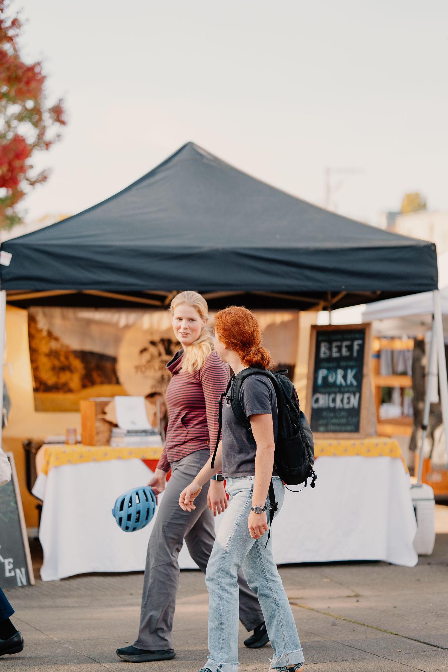 Two people walk past a vendor booth with a black canopy tent at an outdoor market during autumn.
