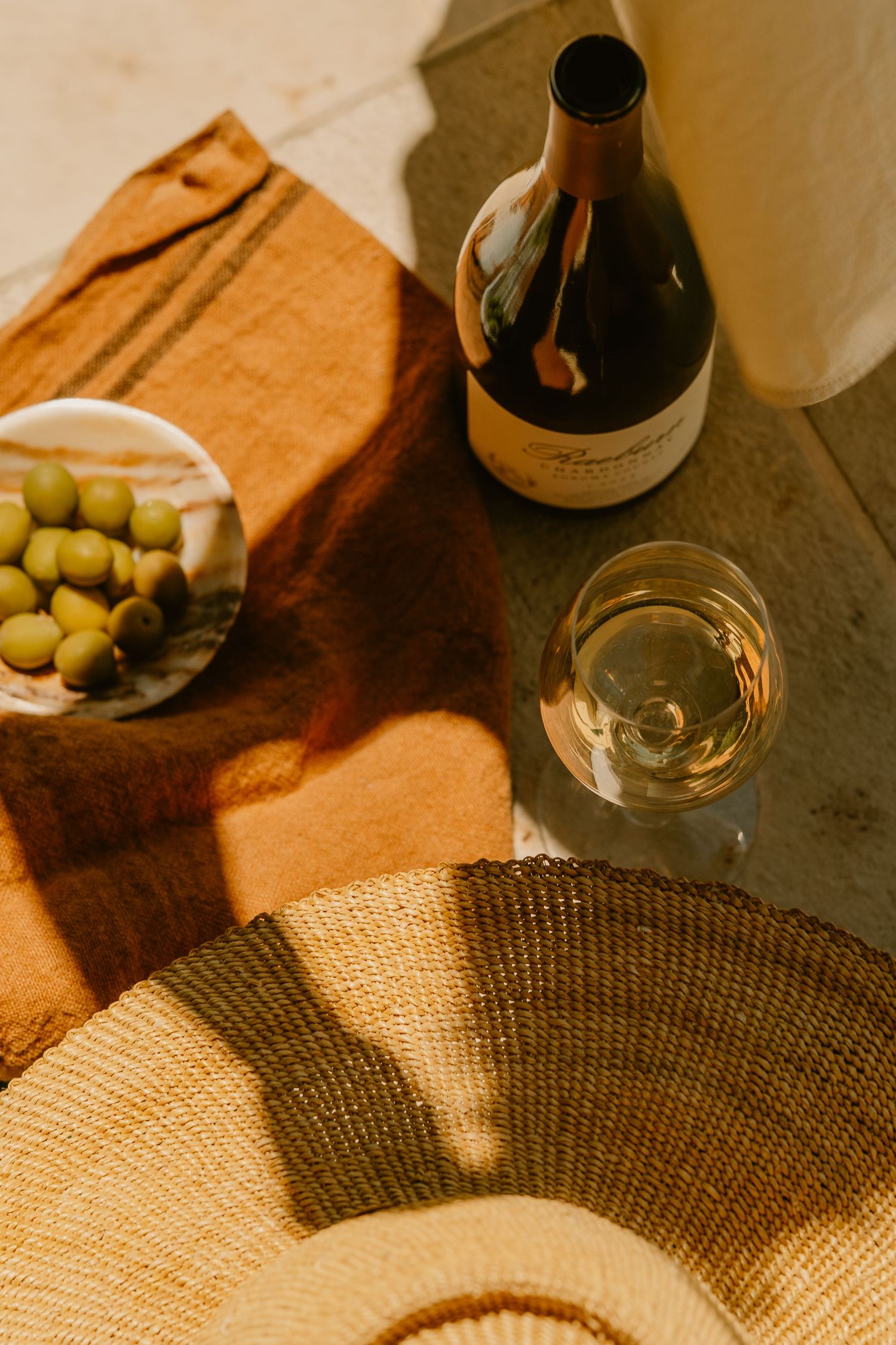 White wine glass and bottle beside a bowl of green olives on a rustic linen cloth with a woven straw hat.