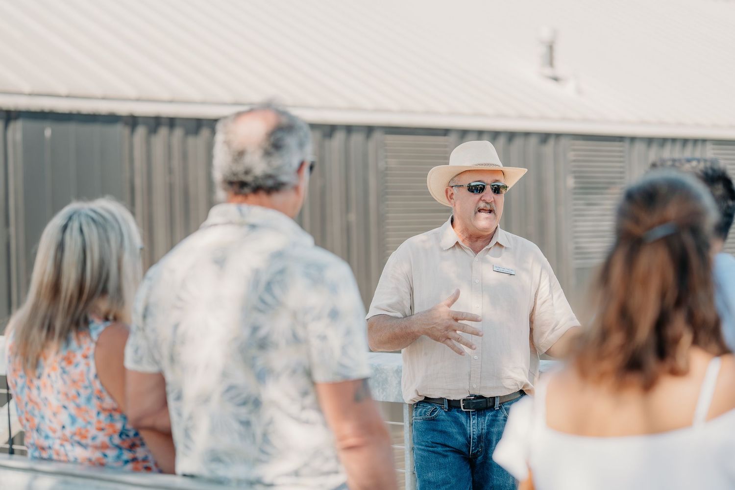 Winery host in cowboy hat and sunglasses speaking to guests during an outdoor vineyard tour.