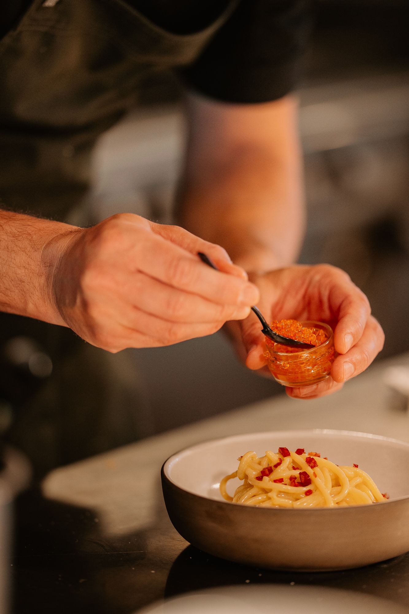 Chef plating spaghetti carbonara with caviar garnish in a white bowl in a professional kitchen.