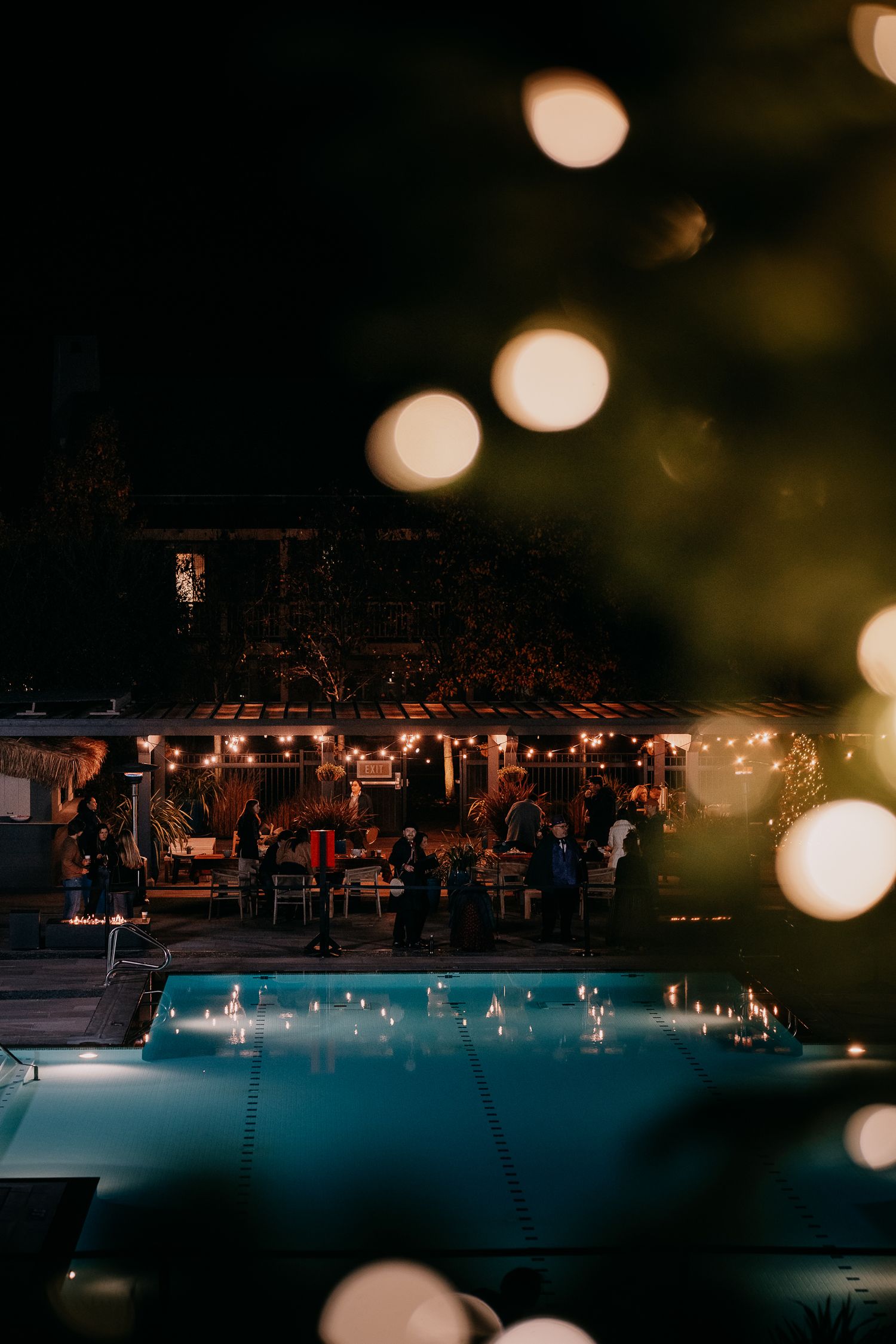Illuminated swimming pool at night with bokeh lights and dark silhouettes creating a moody ambiance.