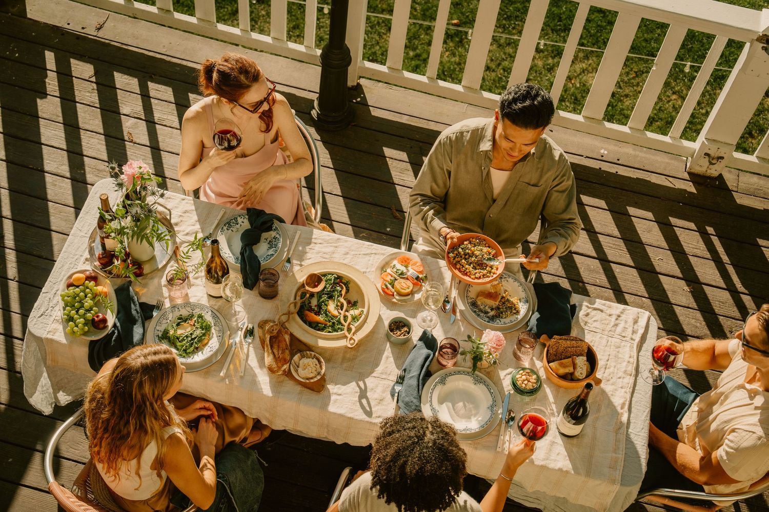 Aerial view of friends enjoying an outdoor dinner party on a sunny deck with fresh salads and food.