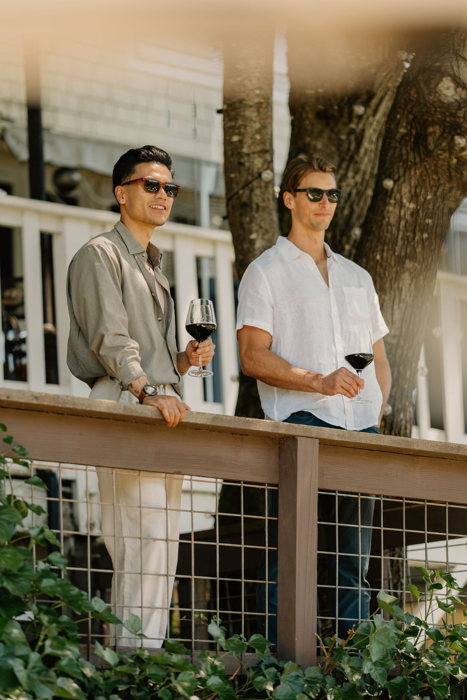 Two men in sunglasses enjoying red wine on a sunny outdoor deck near a white building.