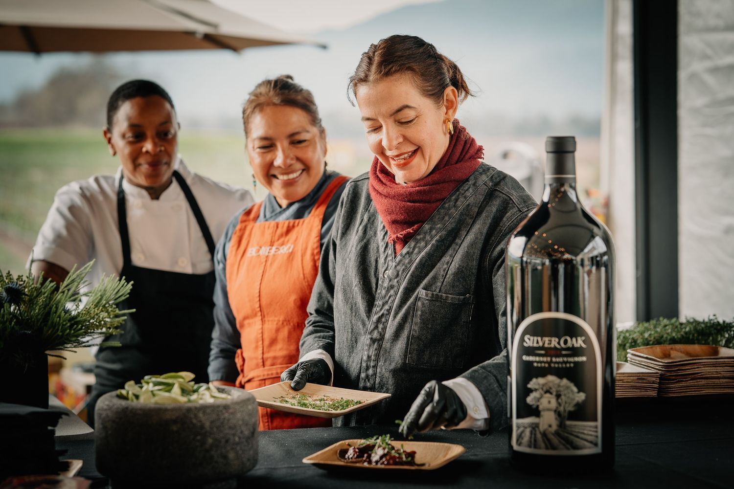 Three women smile while preparing food dishes alongside a Silver Oak wine bottle at culinary event.