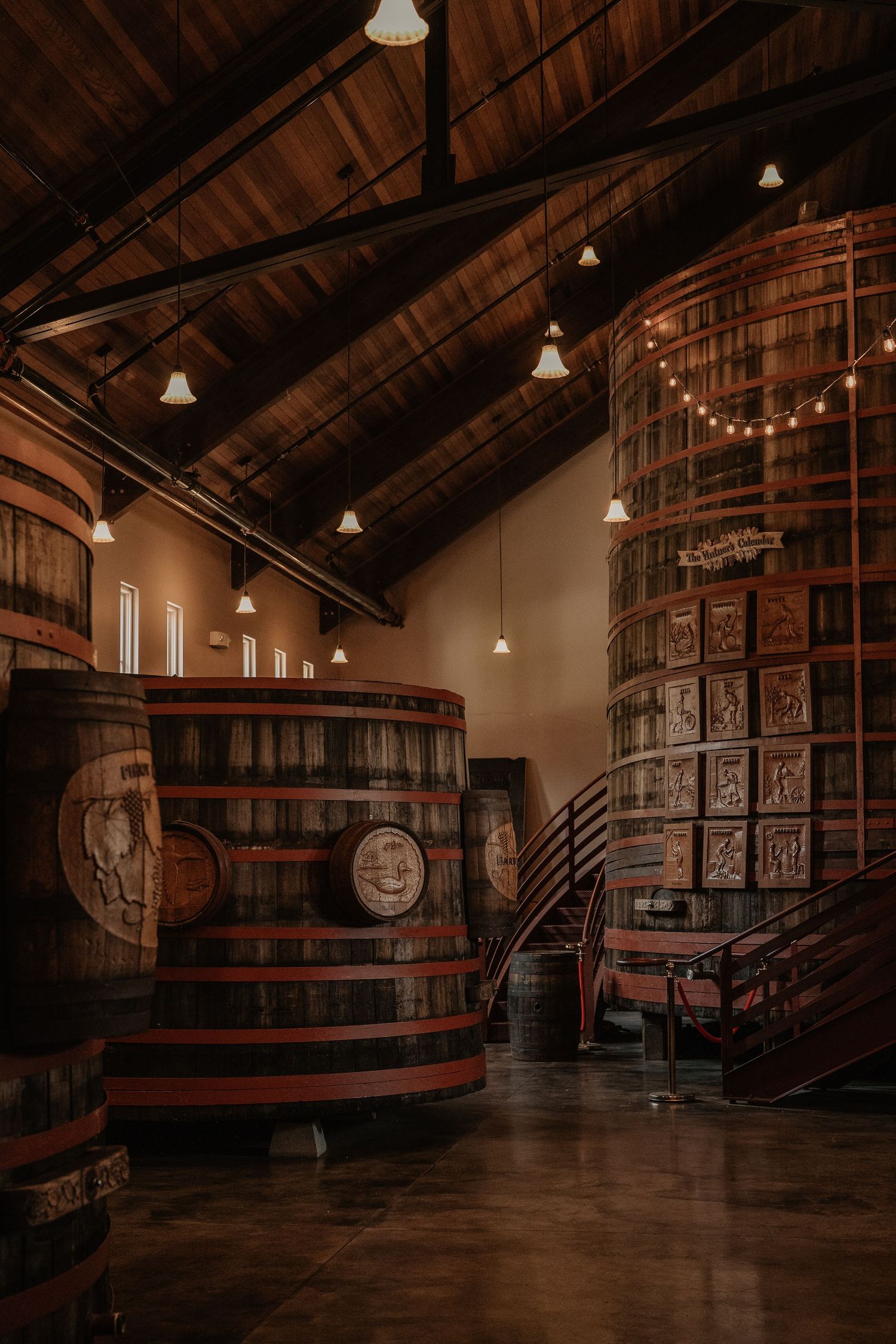 Interior of a rustic winery barrel room with large wooden barrels, warm string lights, and vaulted wooden ceiling.
