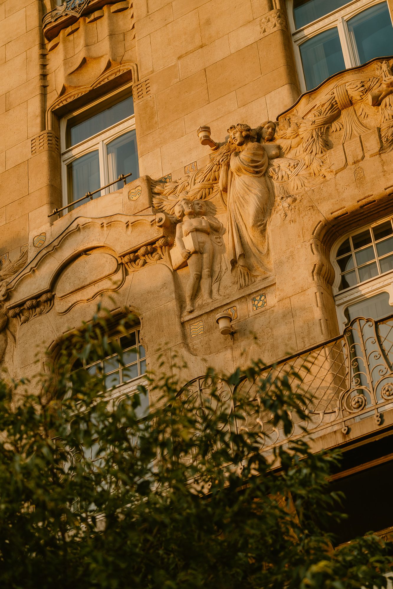 Art Nouveau building facade with ornate stone sculptures of figures bathed in golden sunset light, framed by lush green trees.