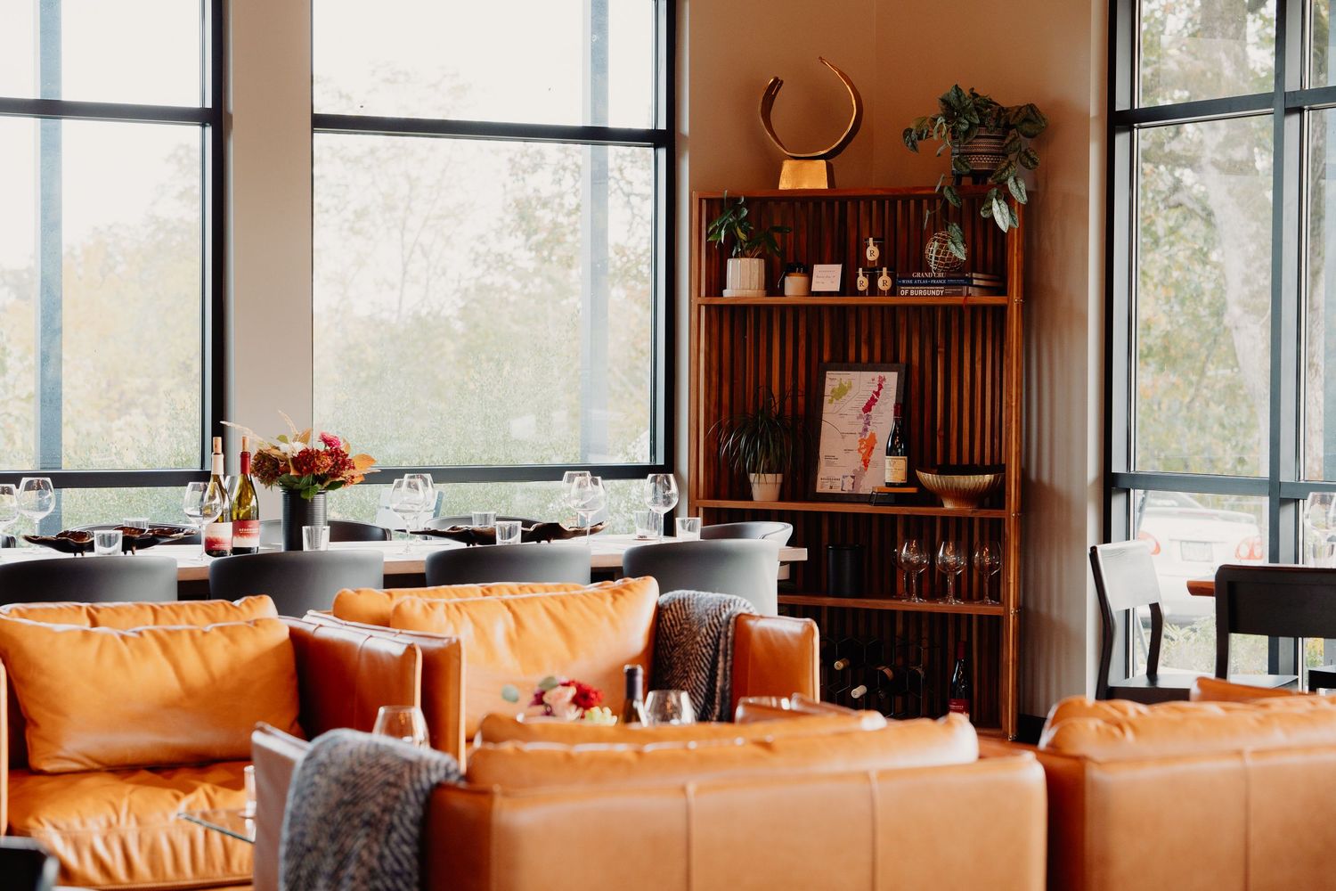 Modern living room interior with tan leather sectional sofa, wood accents, and large windows overlooking natural scenery.