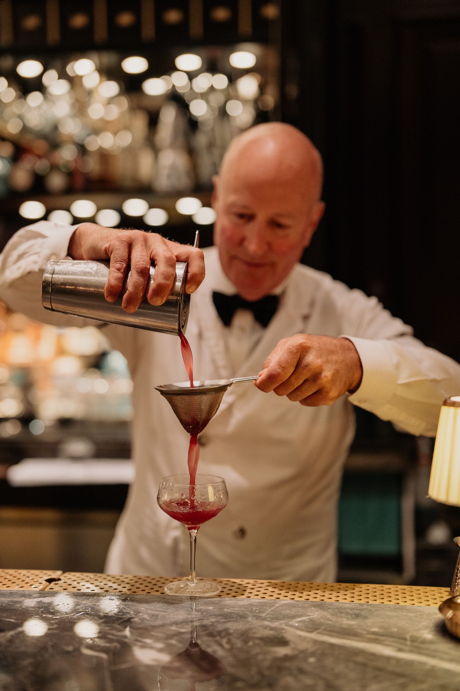 A bartender in a white jacket and bow tie pours a red cocktail into a martini glass at an elegant bar.