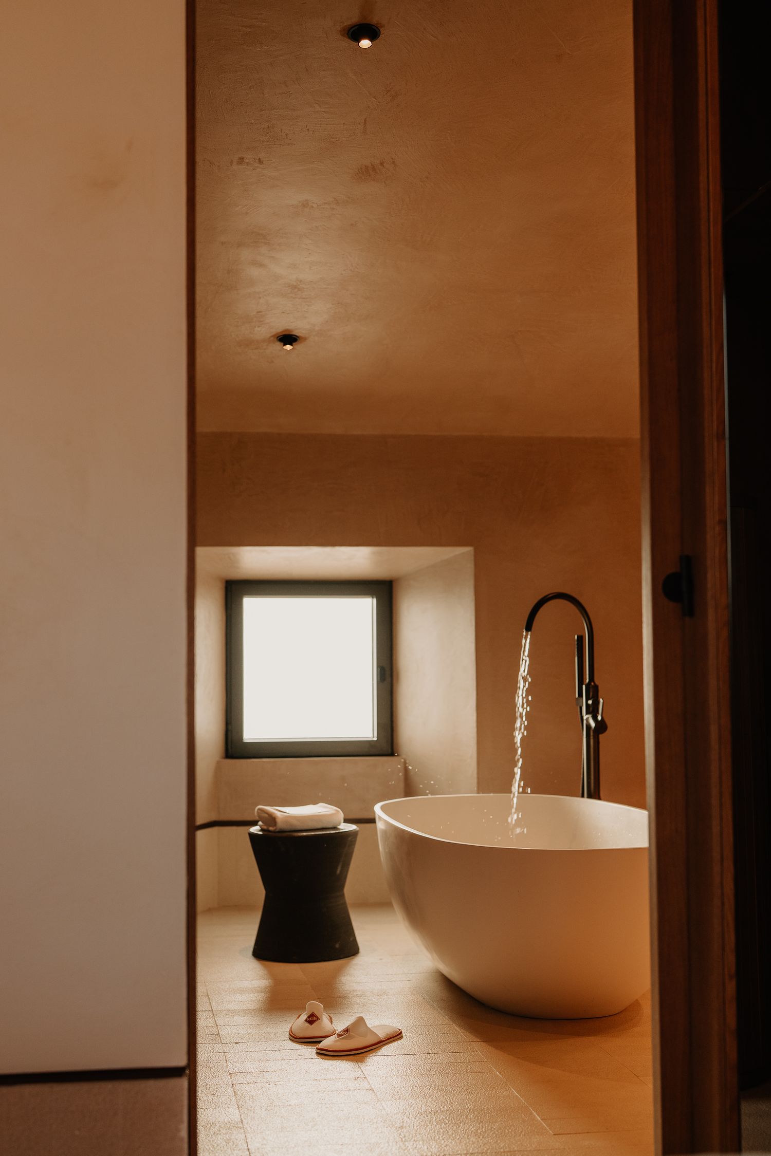 Modern minimalist bathroom interior with freestanding white bathtub, running water, black stool and small window.