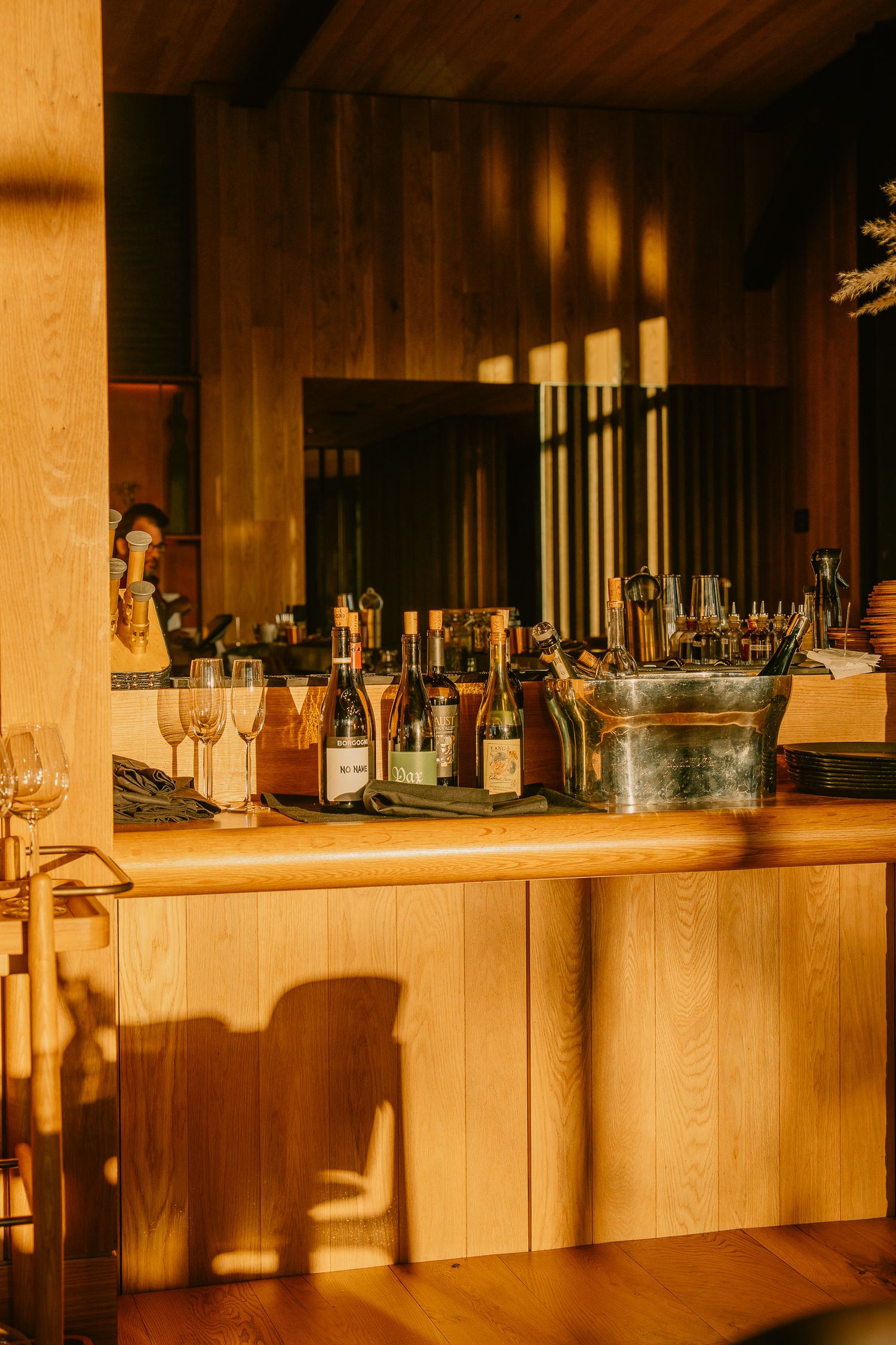 Wooden bar counter with glass bottles and stemware illuminated by natural sunlight.