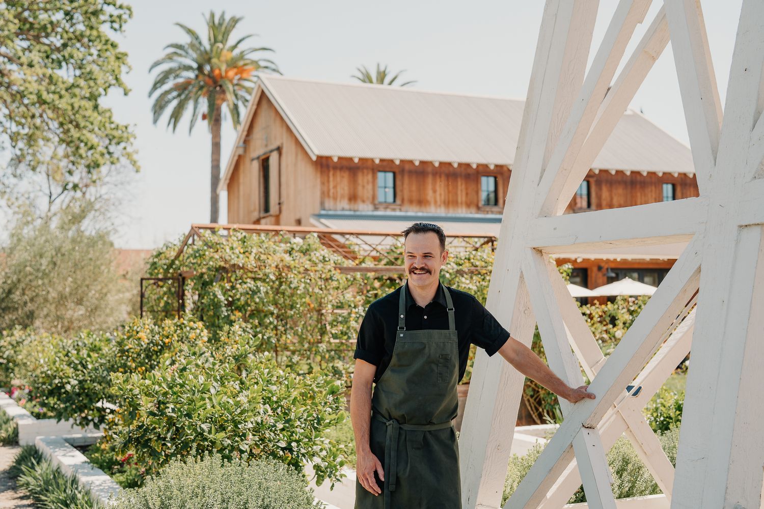 Chef smiling outdoors near a rustic wooden building surrounded by lush greenery and palm trees.
