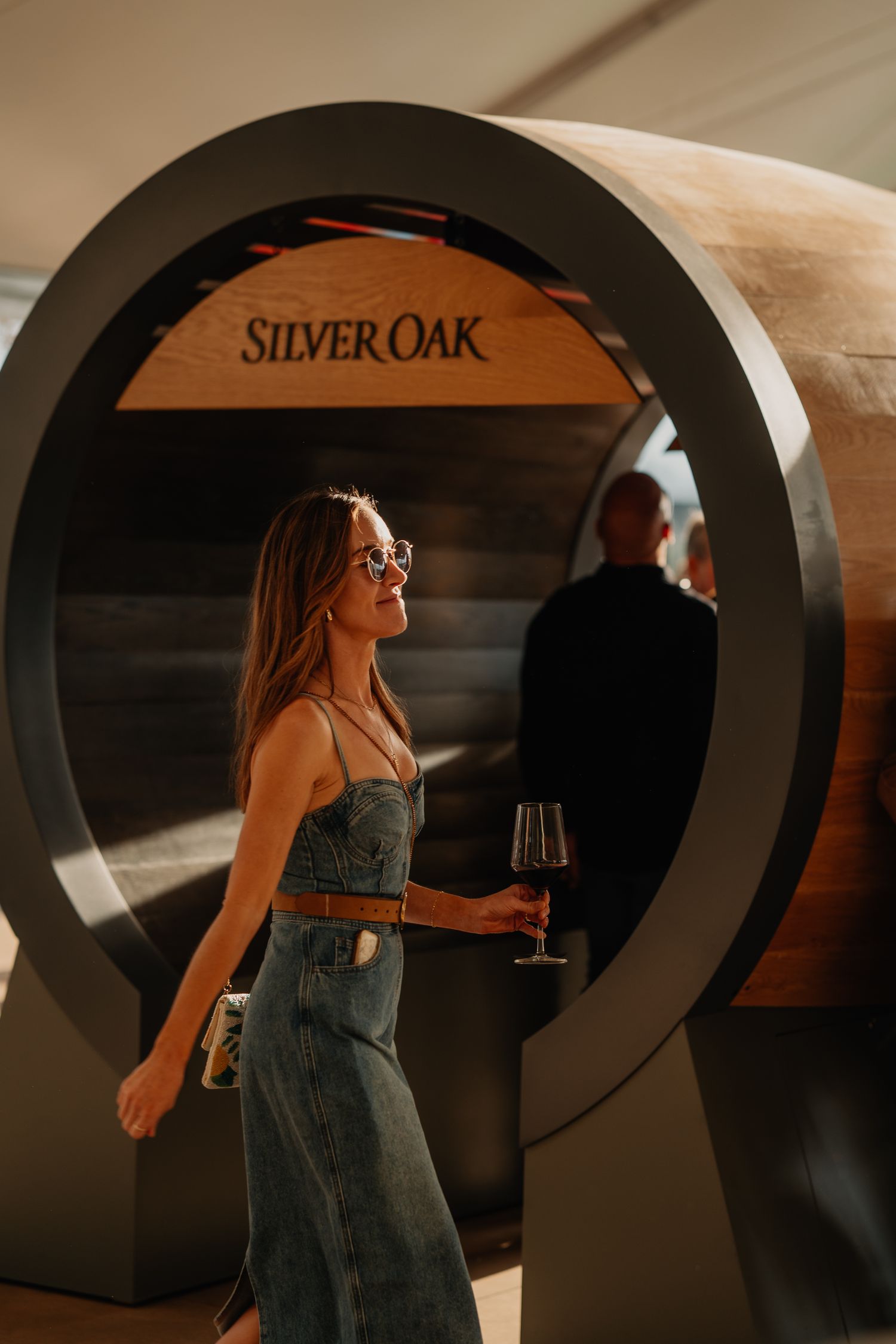 Woman in denim dress holding red wine glass beside a large Silver Oak barrel display installation.