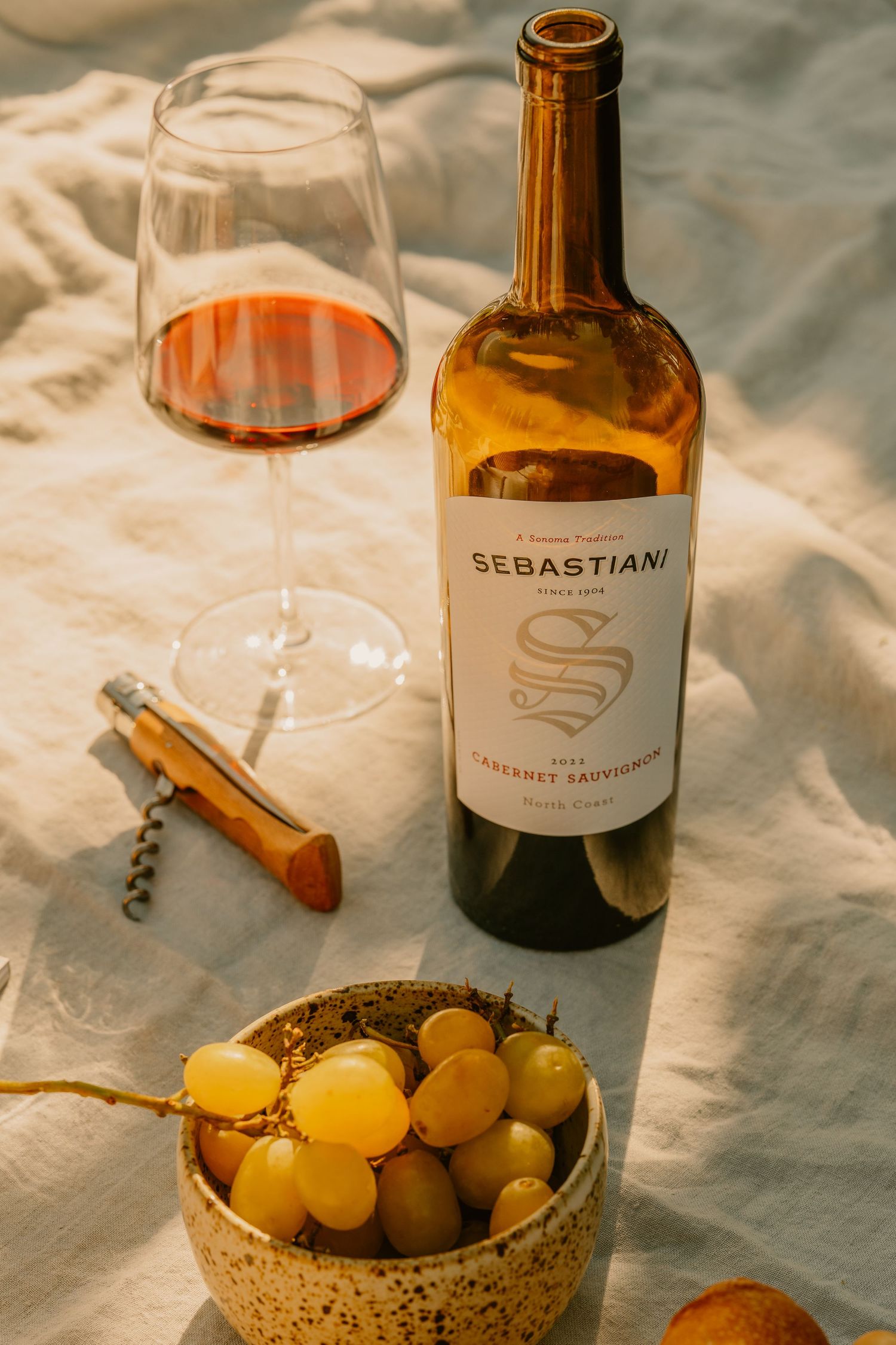 Sebastiani Cabernet Sauvignon bottle with red wine glass, grapes, and corkscrew on white linen cloth.