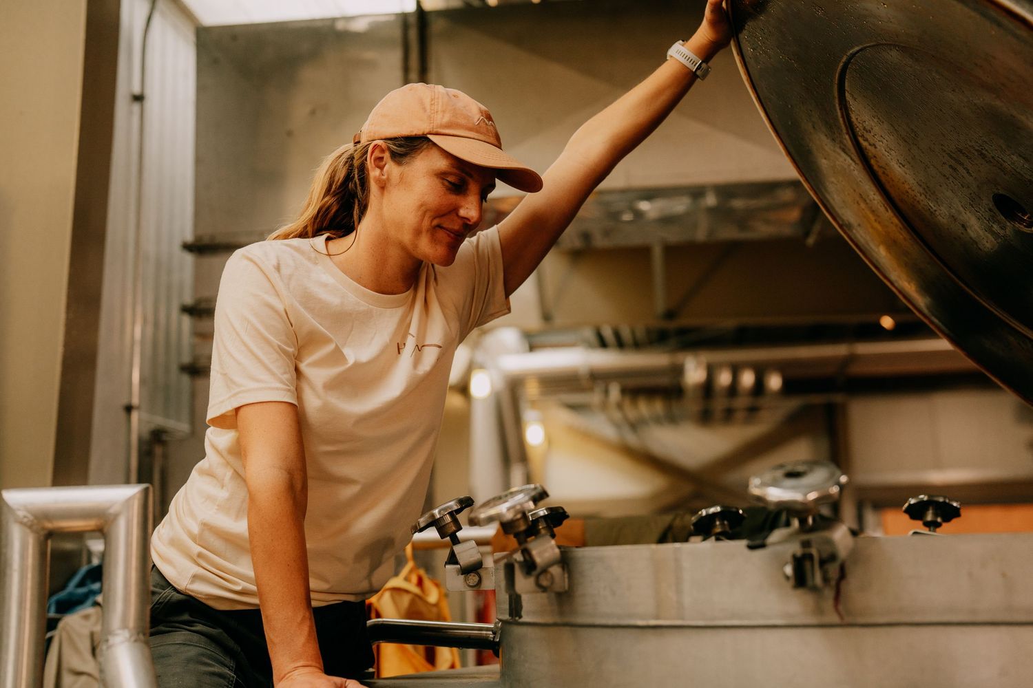 A woman in a cap inspects a large stainless steel fermentation tank in a modern brewery.