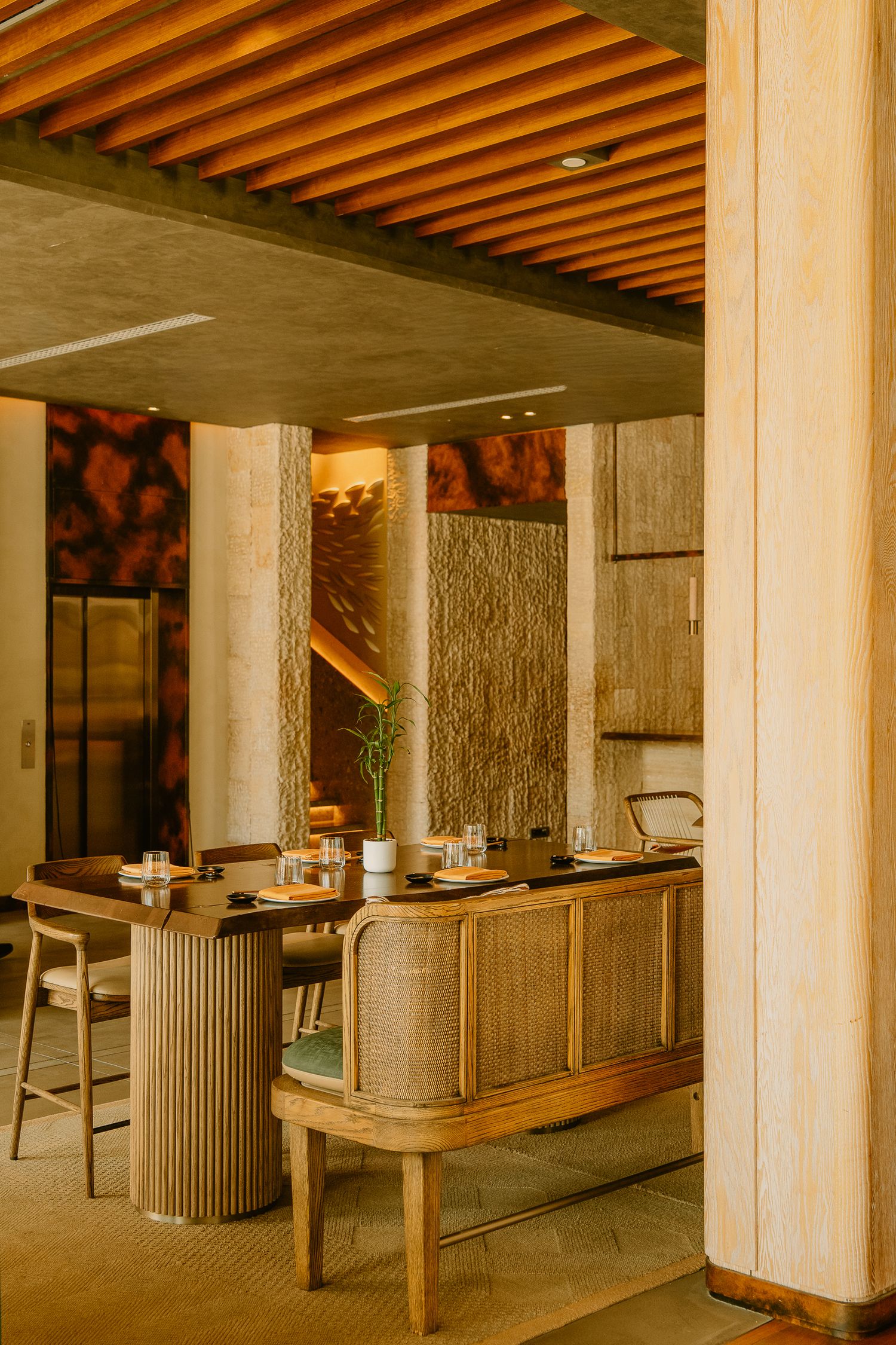 Upscale restaurant interior with wooden slatted ceiling, rattan furniture, and stone columns in warm ambient lighting.