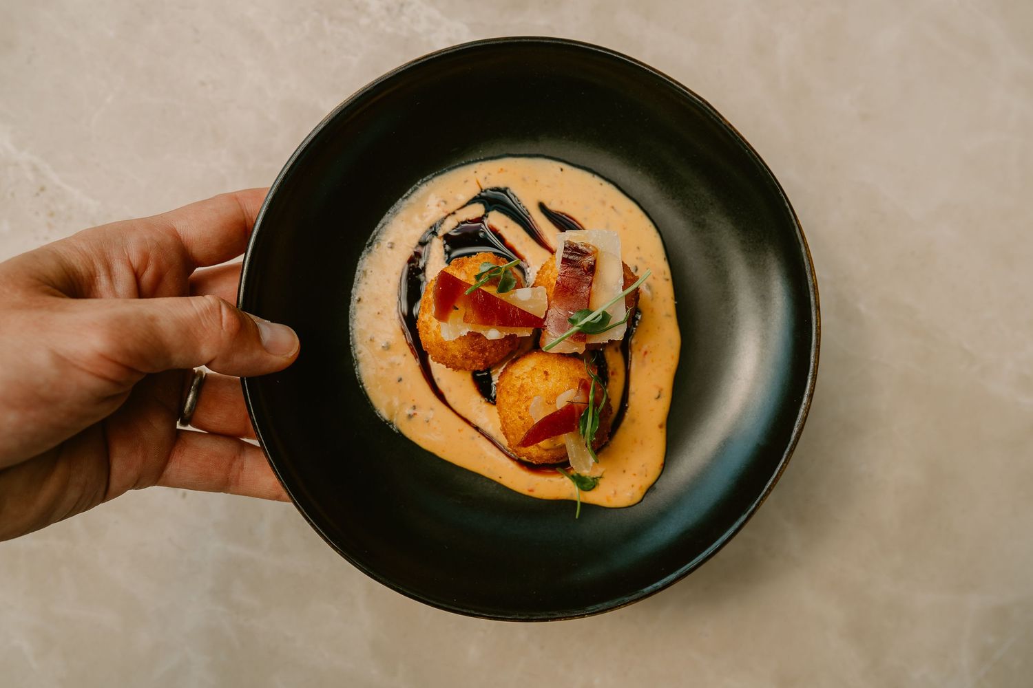 Hand holding a black plate with gourmet dish featuring seared scallops in creamy bisque sauce with garnish.