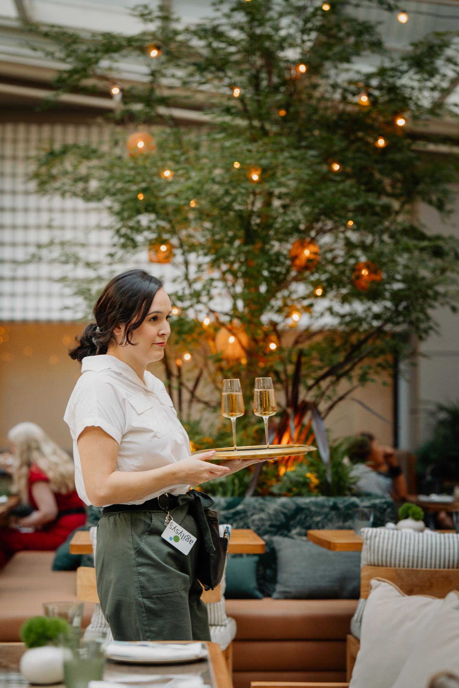 A server carries champagne flutes through an elegant outdoor dining area with string lights and greenery.