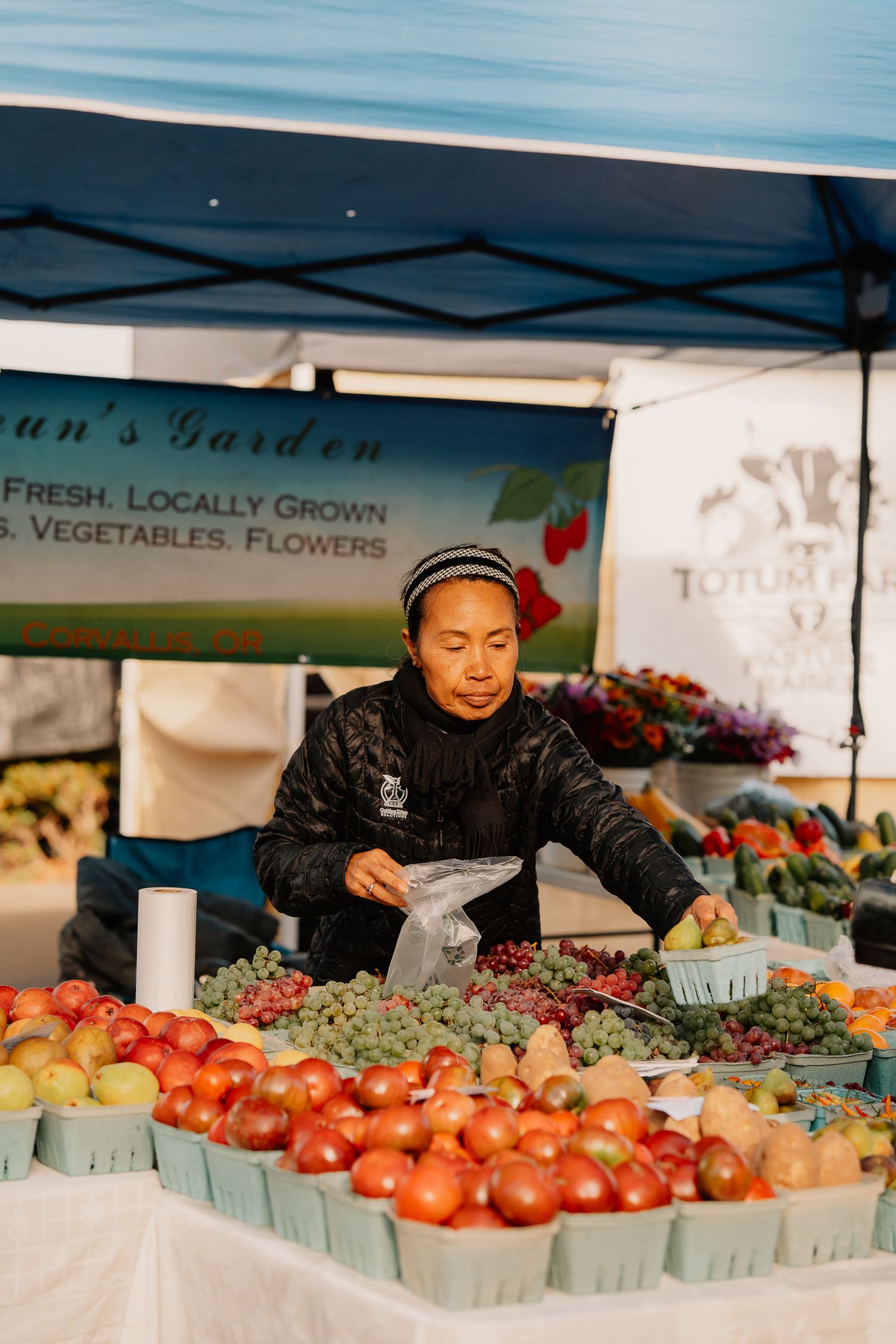 Fresh produce vendor at farmers market displays colorful tomatoes and other vegetables under a garden stand tent.