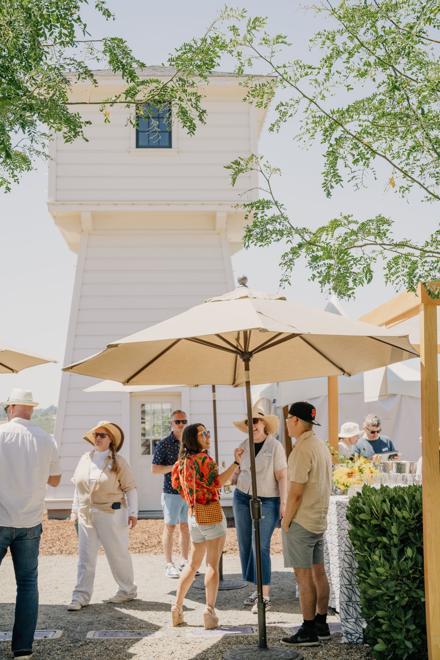 Guests socializing under tan patio umbrellas at an outdoor event near a white lighthouse building on a sunny day.