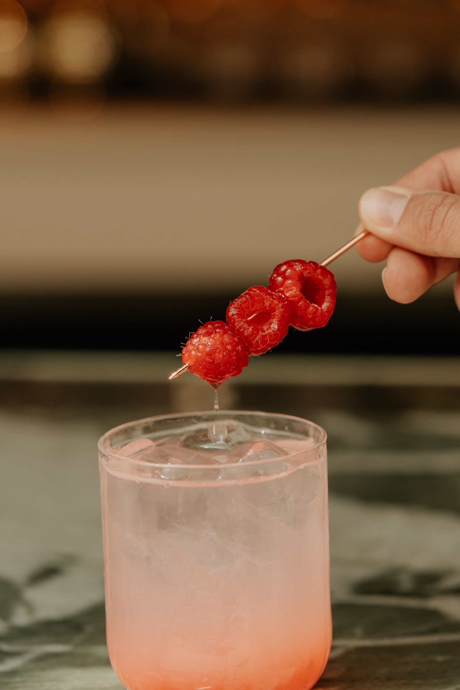 Hand garnishing a pink cocktail with fresh red raspberries on a wooden skewer over a frosted glass.
