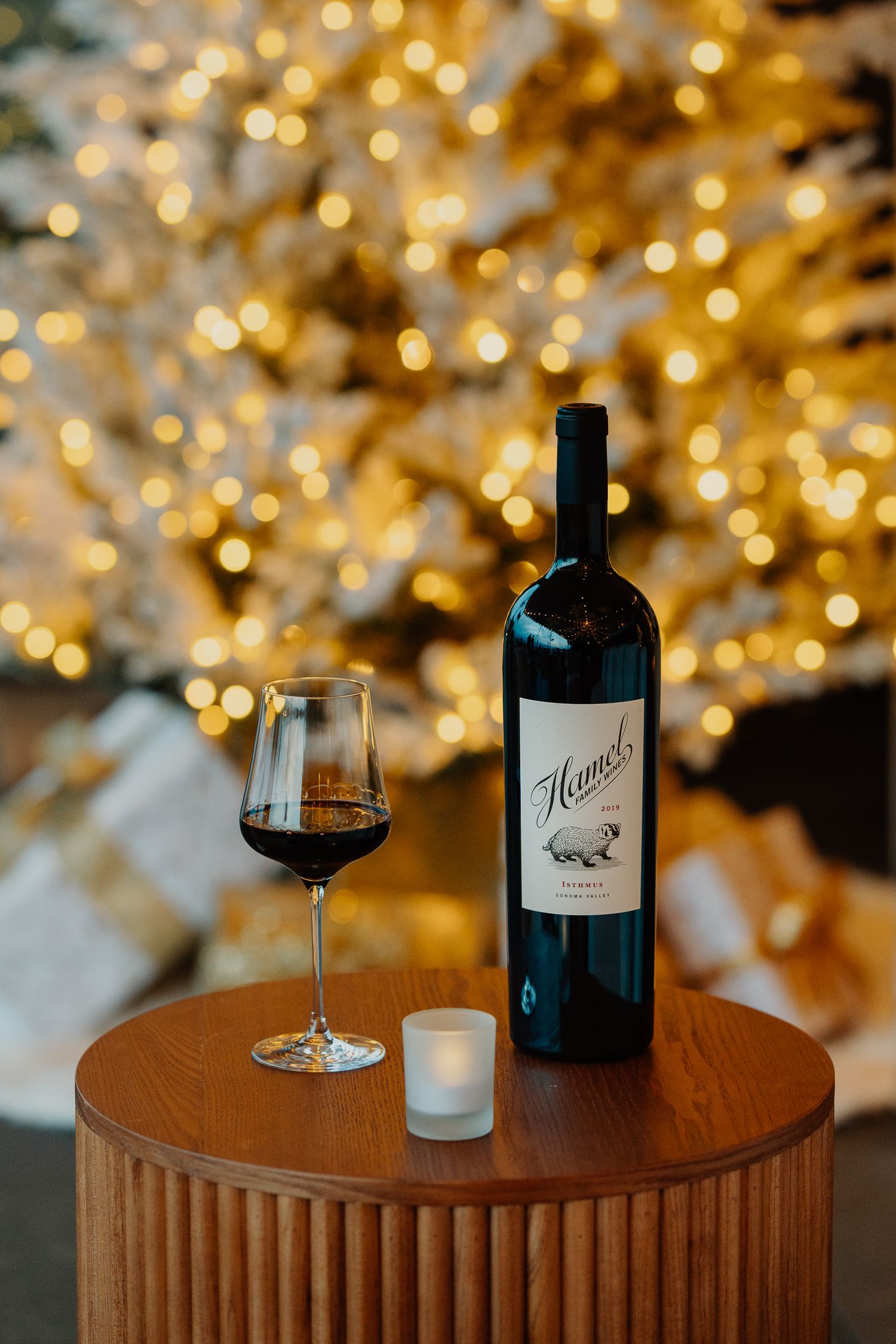 A glass of red wine and wine bottles displayed on a wooden table with soft glowing bokeh lights in the background.