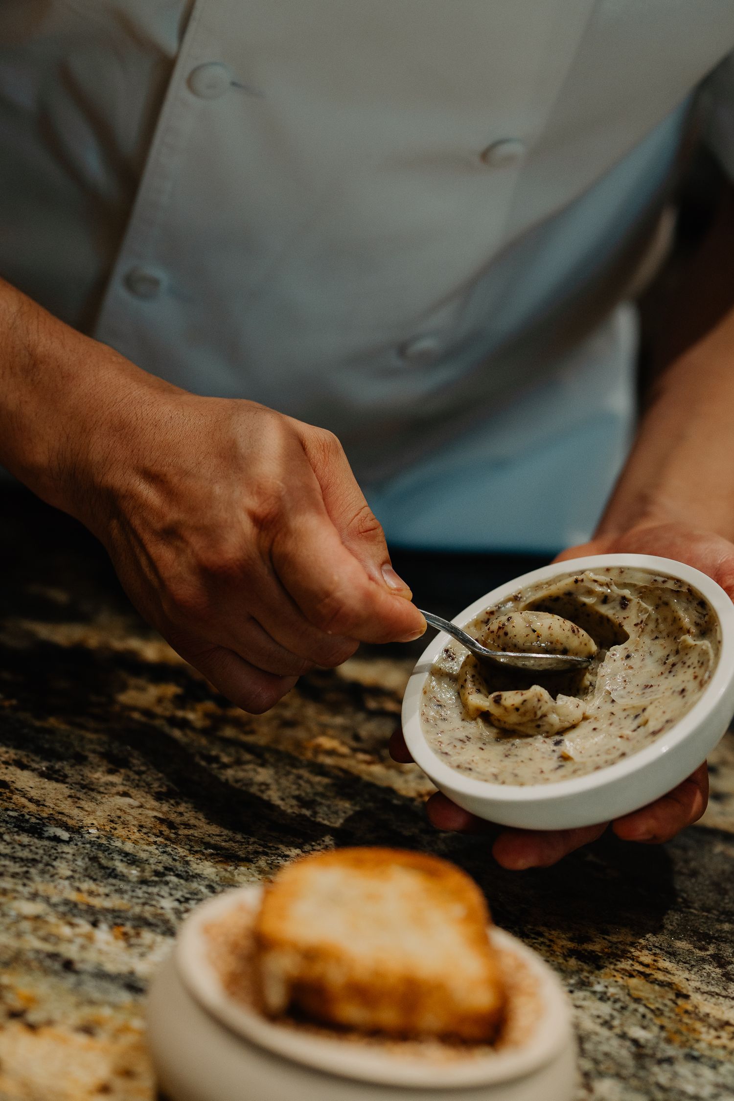 Chef in white uniform scoops creamy spread onto rustic marble counter with toasted bread in foreground.