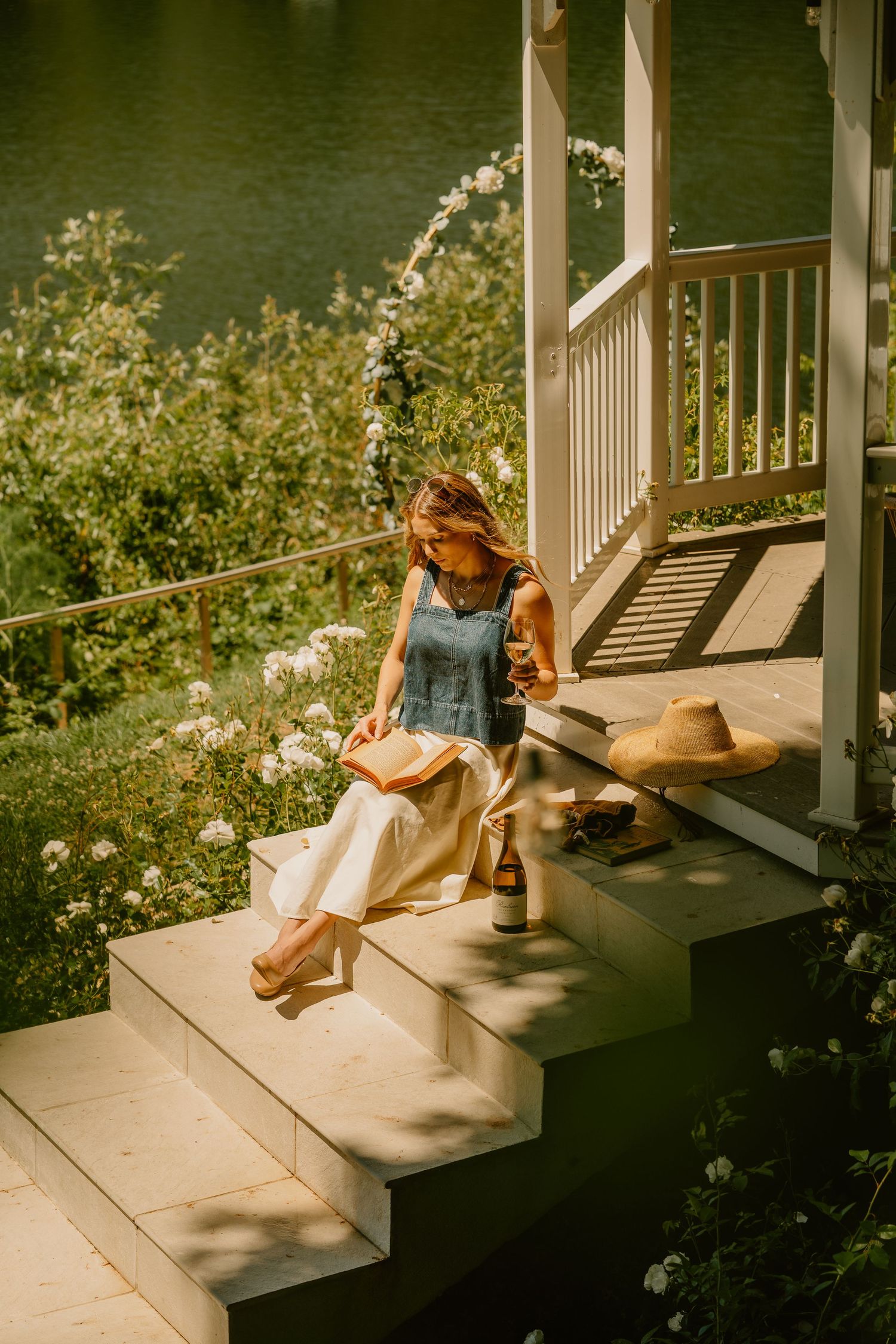 Woman reading a book on sunlit porch steps beside a straw hat and bottle, surrounded by blooming flowers near a lake.