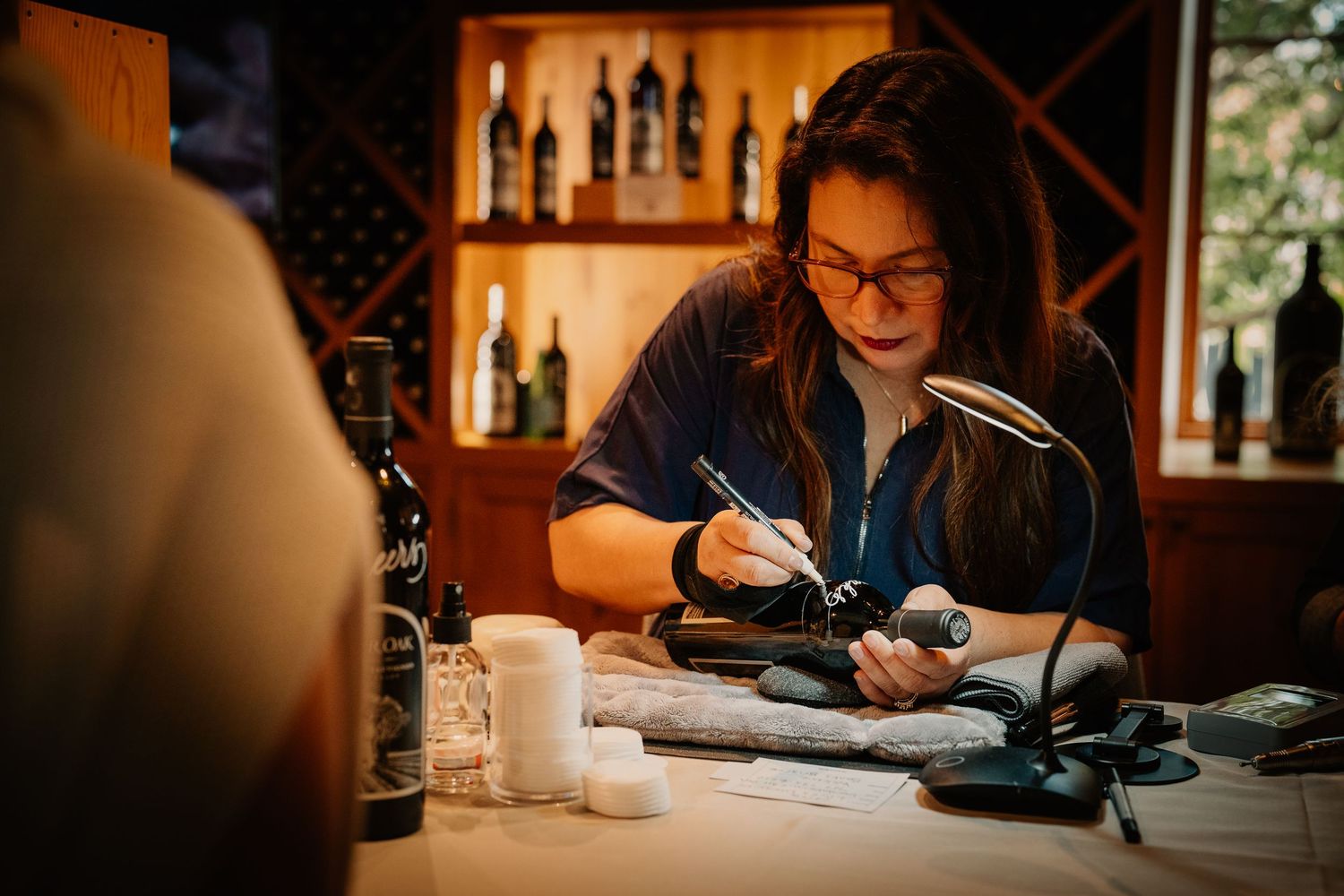 A tattoo artist carefully working on a design at a table in a warmly lit wine bar setting.