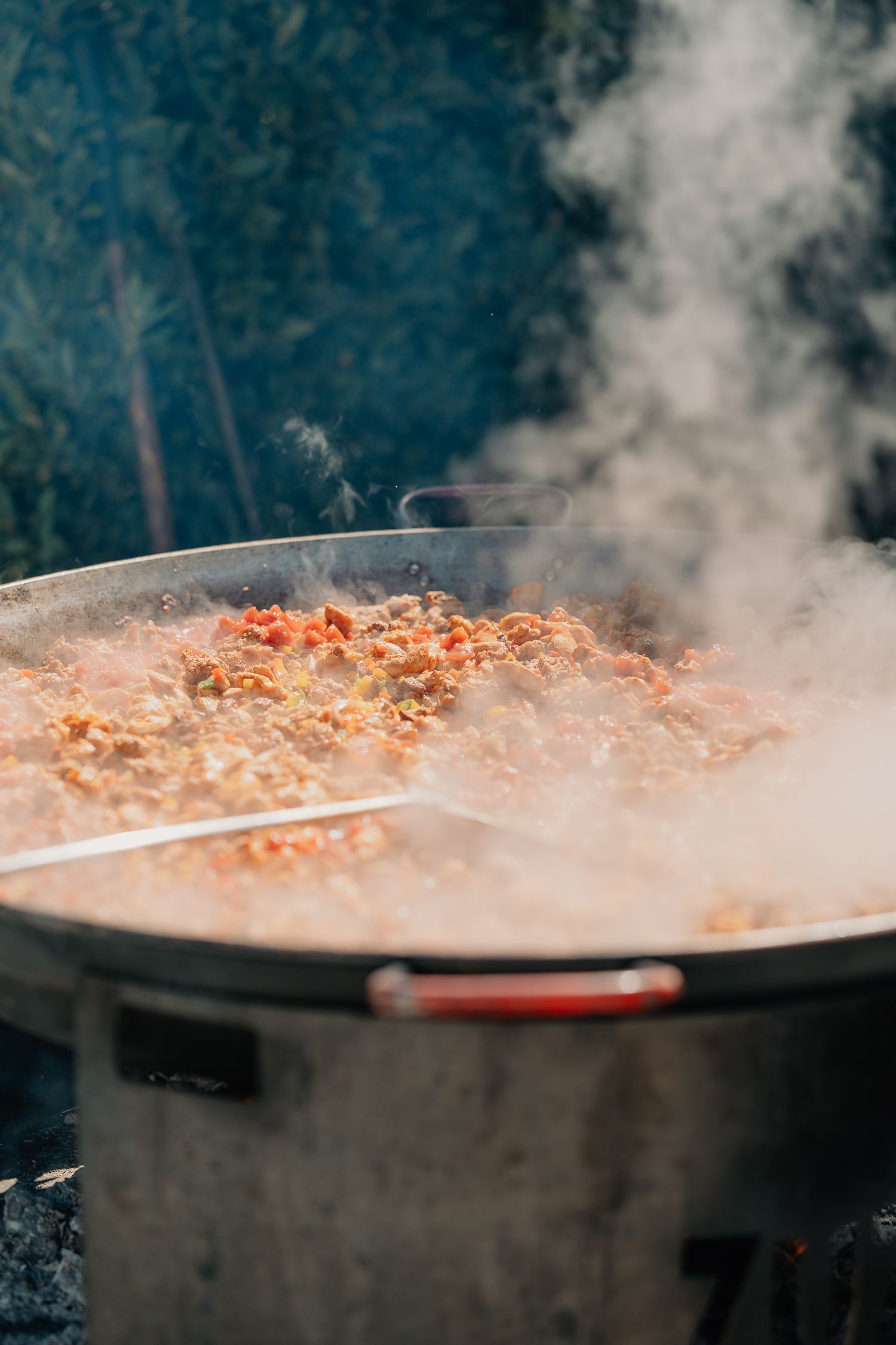 Steaming large paella pan filled with rice, vegetables, and meat cooking outdoors over an open flame.