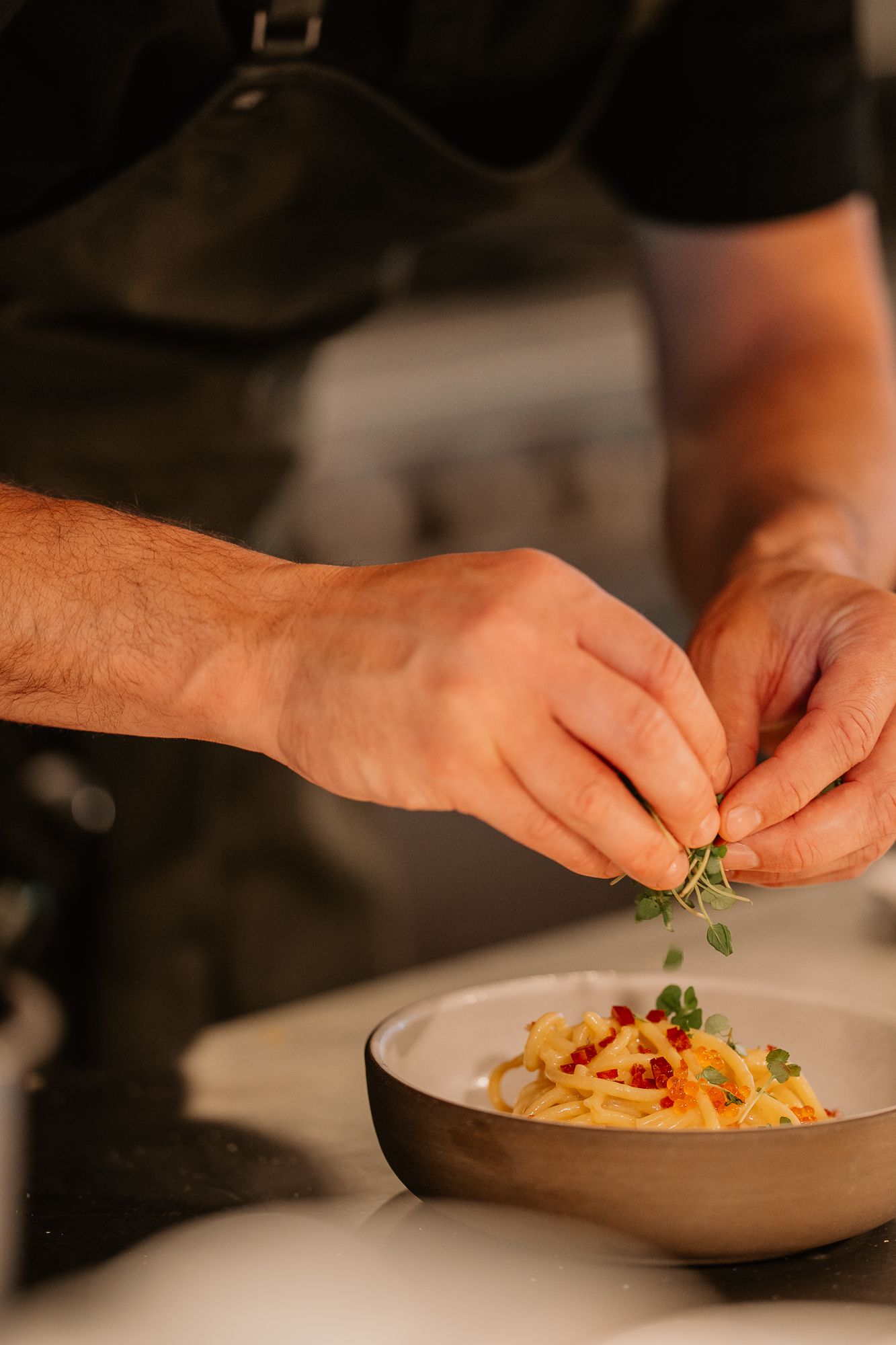 Chef garnishing a bowl of spaghetti carbonara with fresh herbs in a professional kitchen.