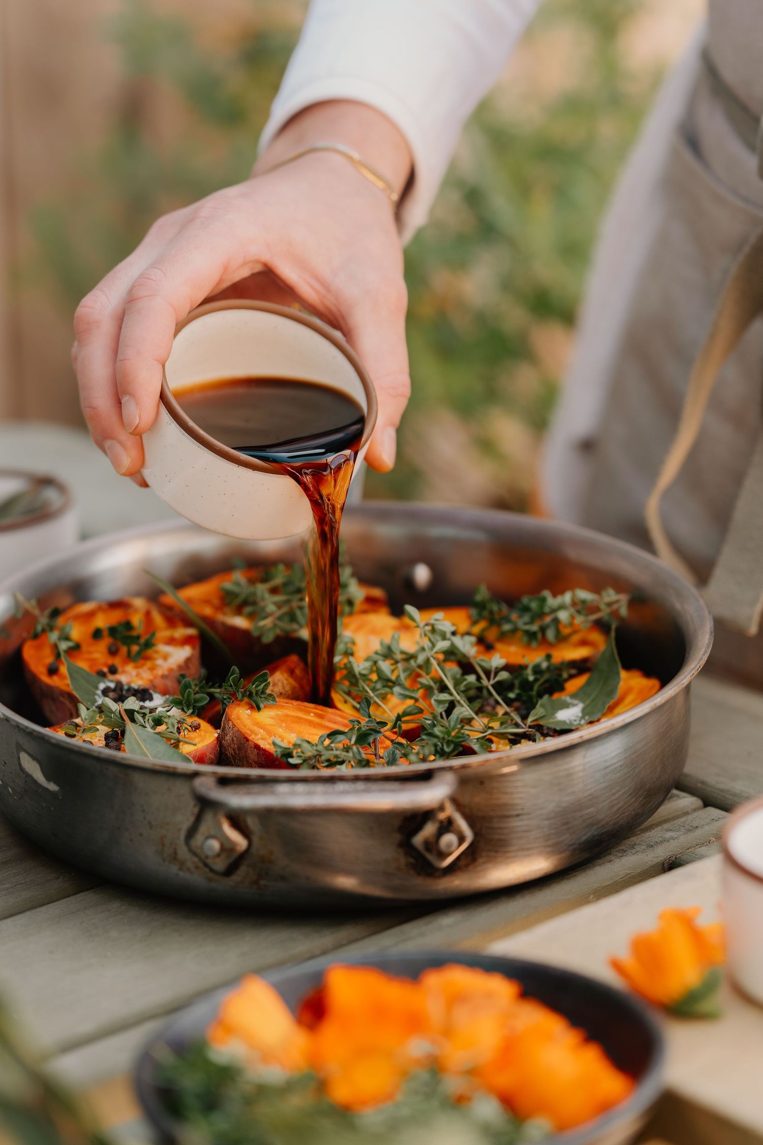 Hand pouring dark sauce over roasted pumpkin slices garnished with fresh thyme in a pan.