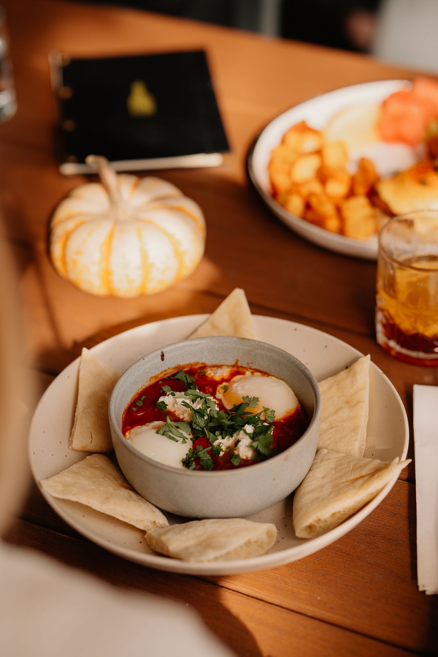 A cozy table setting with queso dip, tortilla chips, decorative white pumpkin, and a cocktail in warm lighting.