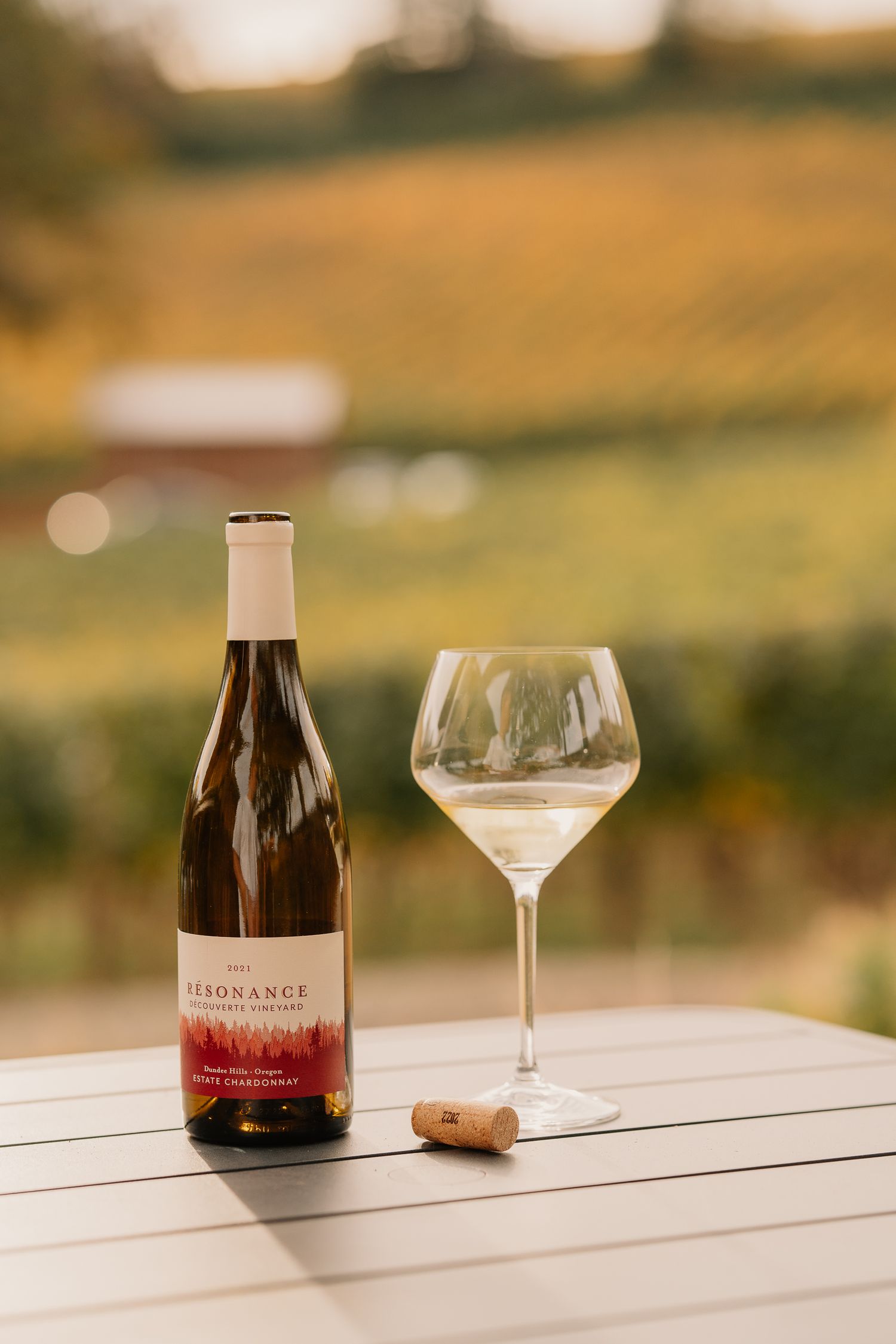 A bottle of Resonance wine and glass of white wine sit on wooden table with vineyard landscape in background.